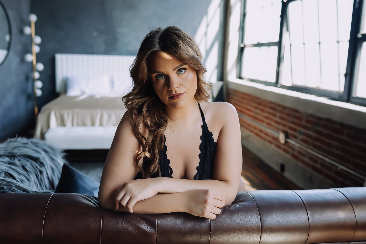 Woman in black lingerie posing on a leather sofa in a stylish loft bedroom with brick walls.