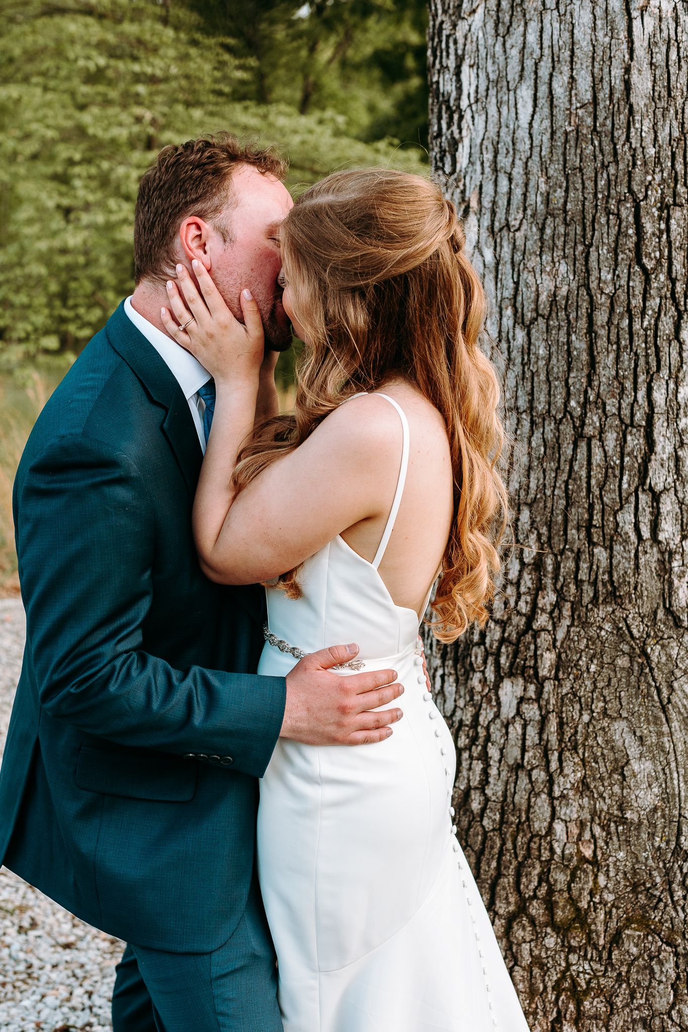 Romantic wedding couple kiss against large tree trunk in elegant attire.