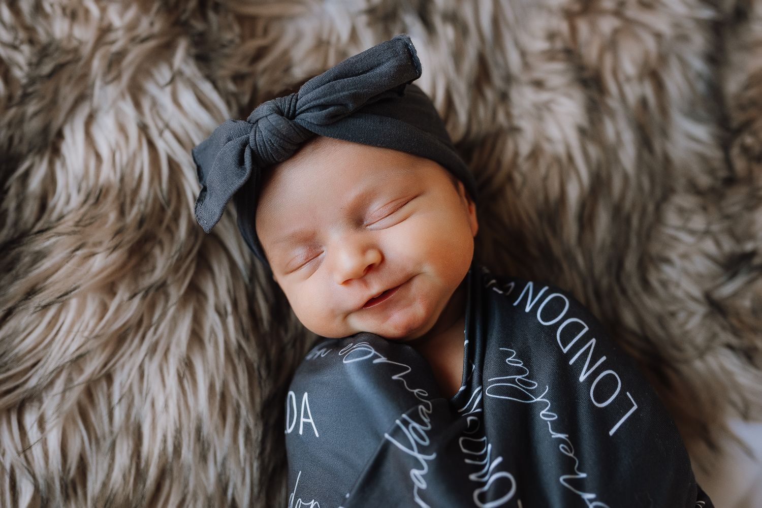 Smiling newborn baby swaddled in a dark gray personalized blanket with a matching bow headband on a fluffy rug.