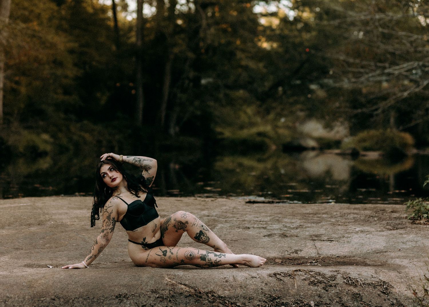 Heavily tattooed woman posing on rocky ground near a forest creek wearing black bikini.