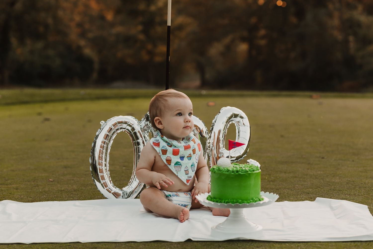 Baby sits on white blanket with green birthday cake and silver number balloons during sunset photoshoot.