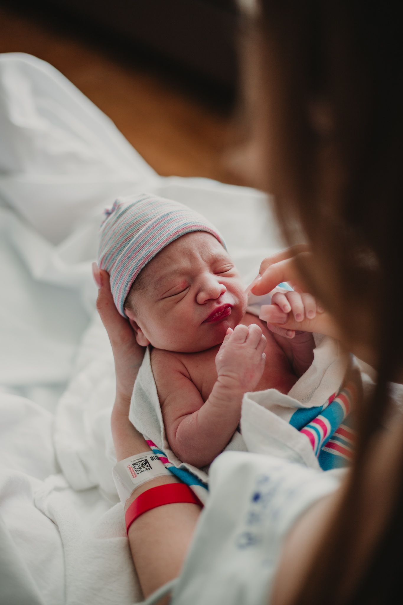 Newborn baby in striped hat held by mother in hospital, gripping her finger with tiny hand after birth.