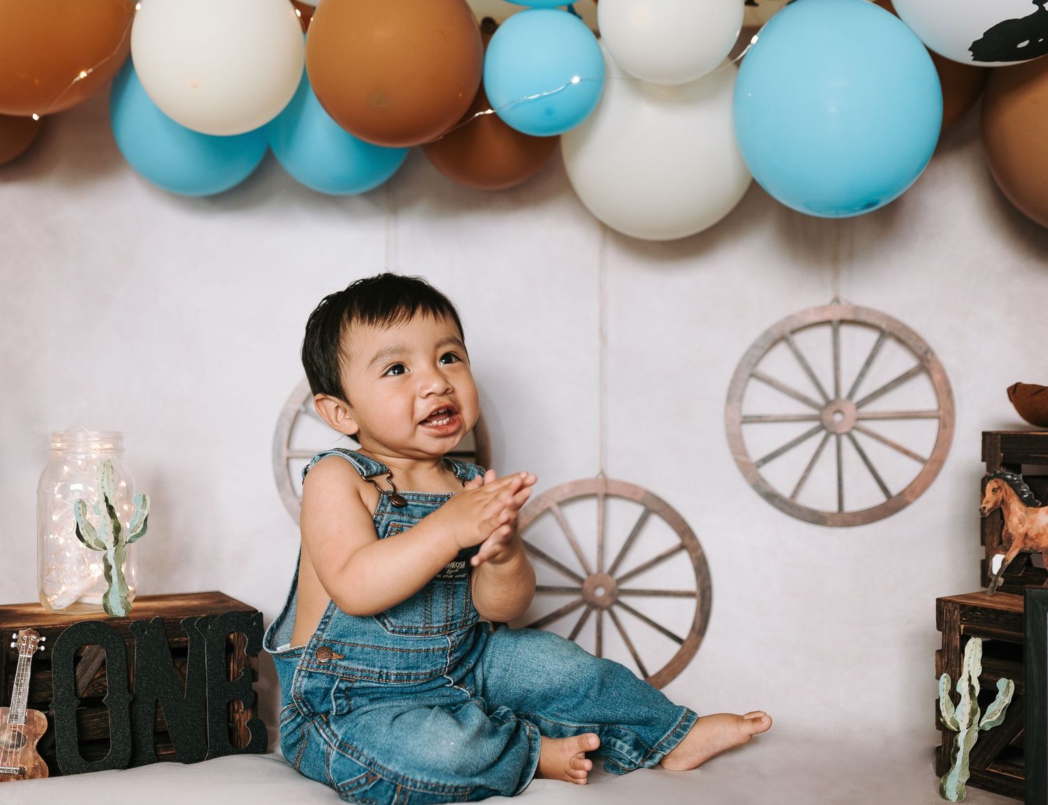 Young child in denim overalls sits on floor with western-themed party decorations and blue balloons.
