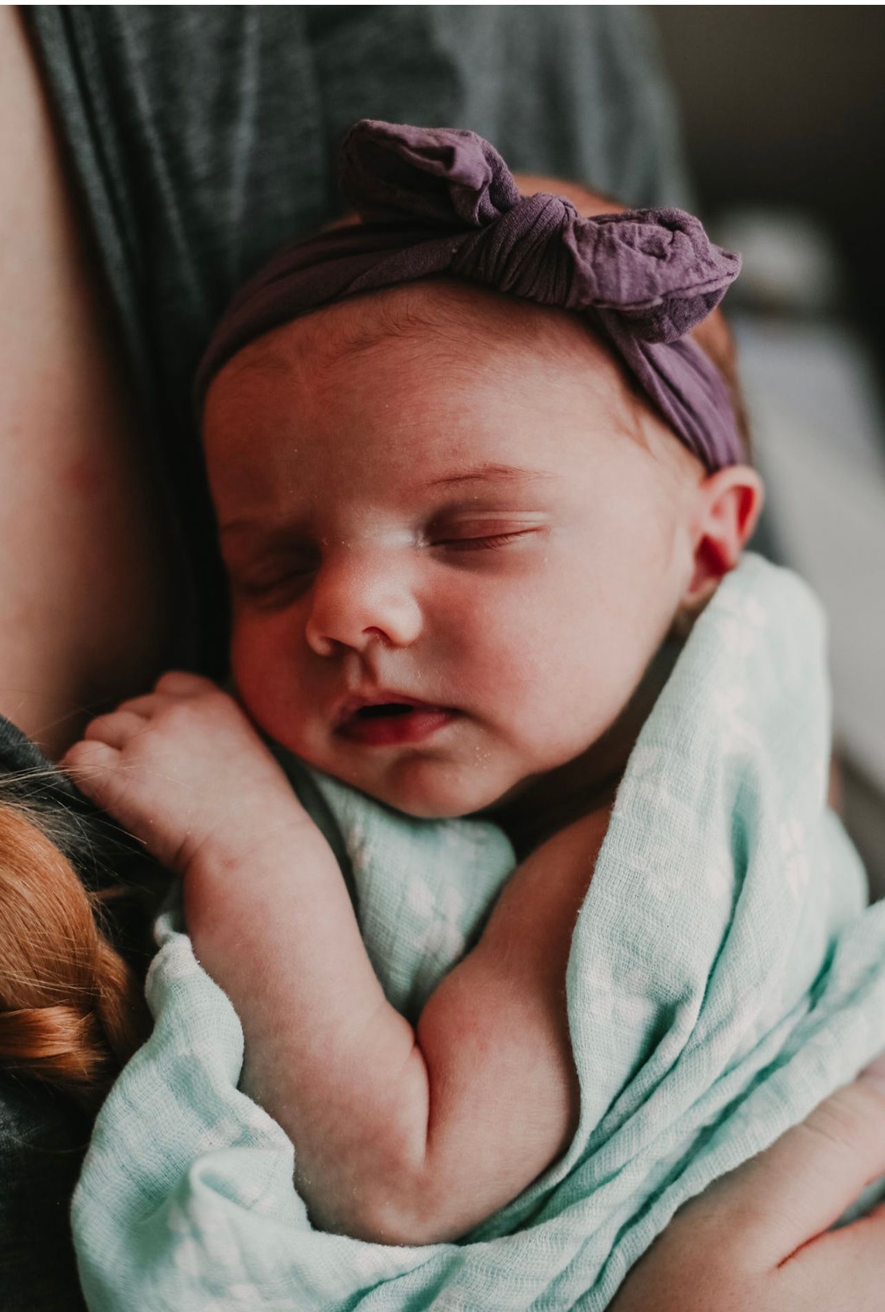 A sleeping newborn wearing a purple headband rests peacefully in mint green swaddle.
