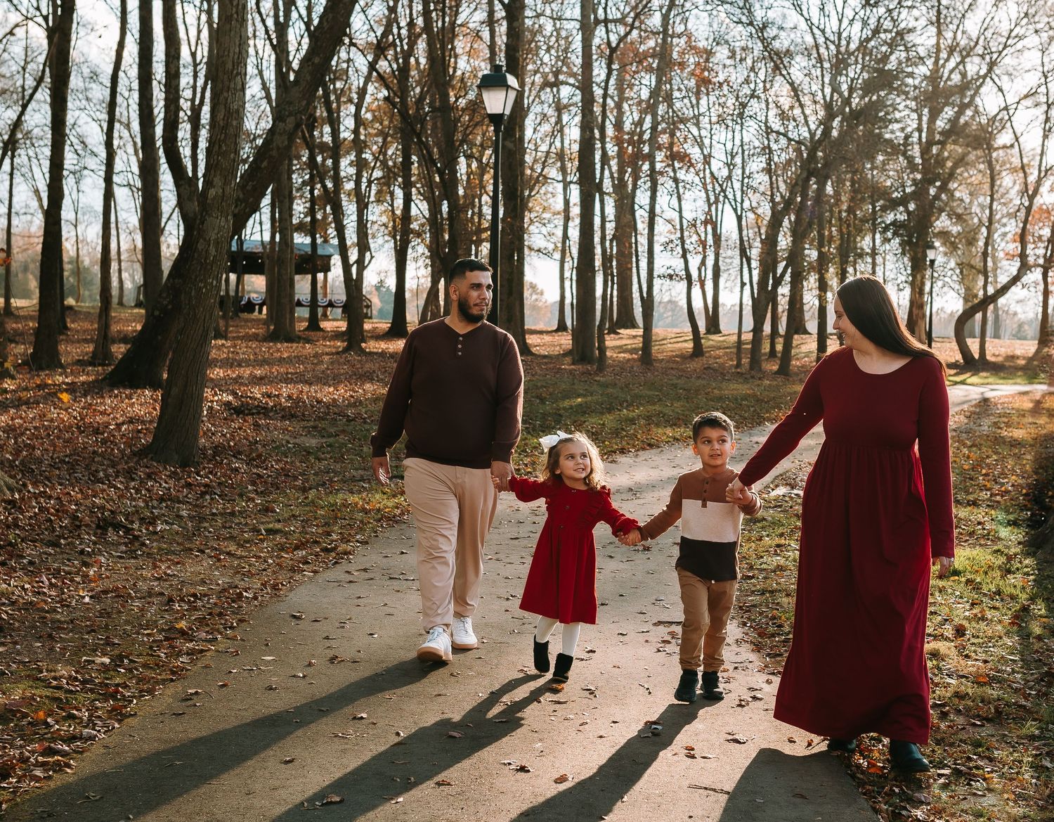 Family of four walking hand-in-hand on a park path surrounded by autumn trees and fallen leaves.