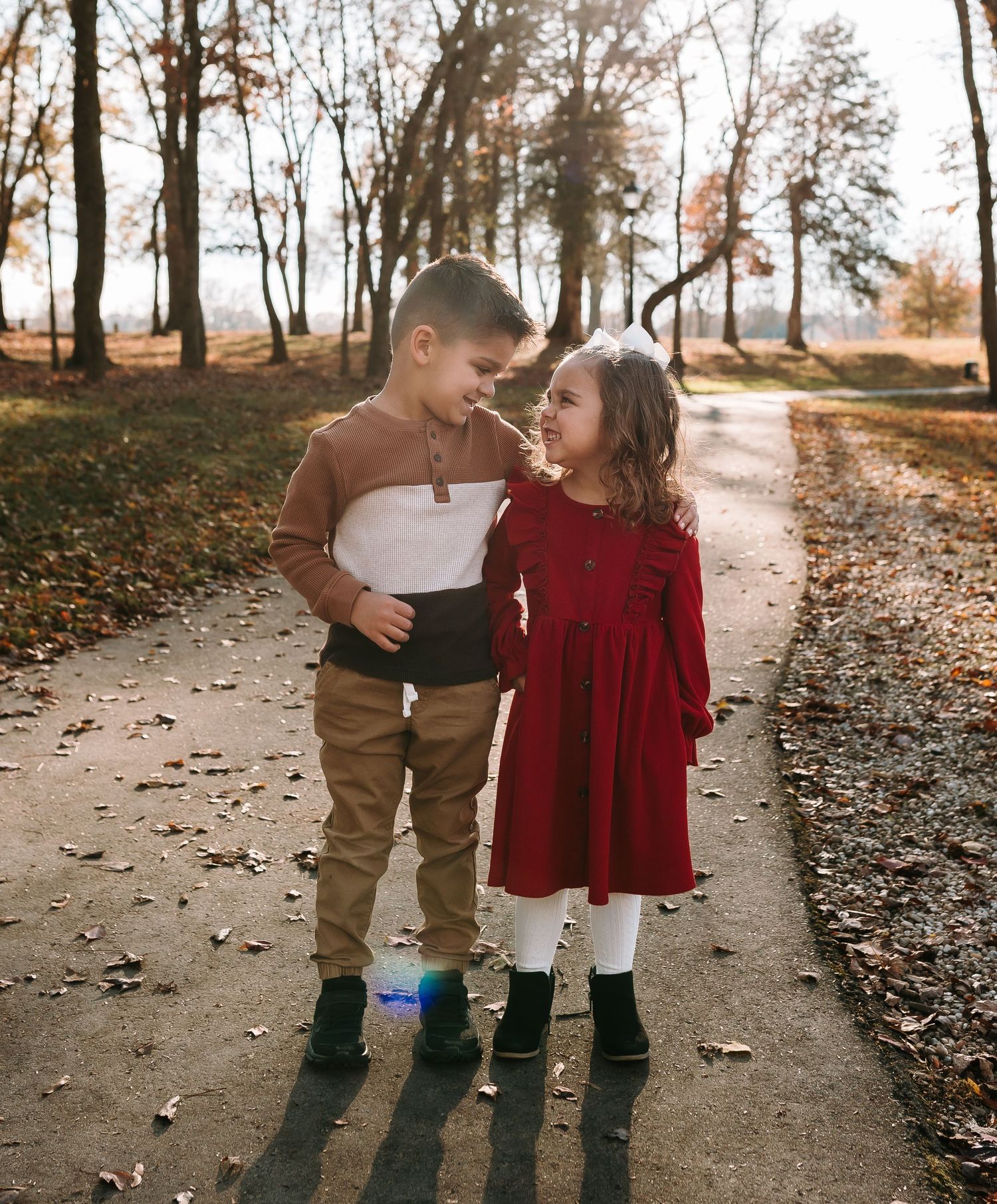 Two young children in autumn outfits smile at each other on a tree-lined path covered in fallen leaves.
