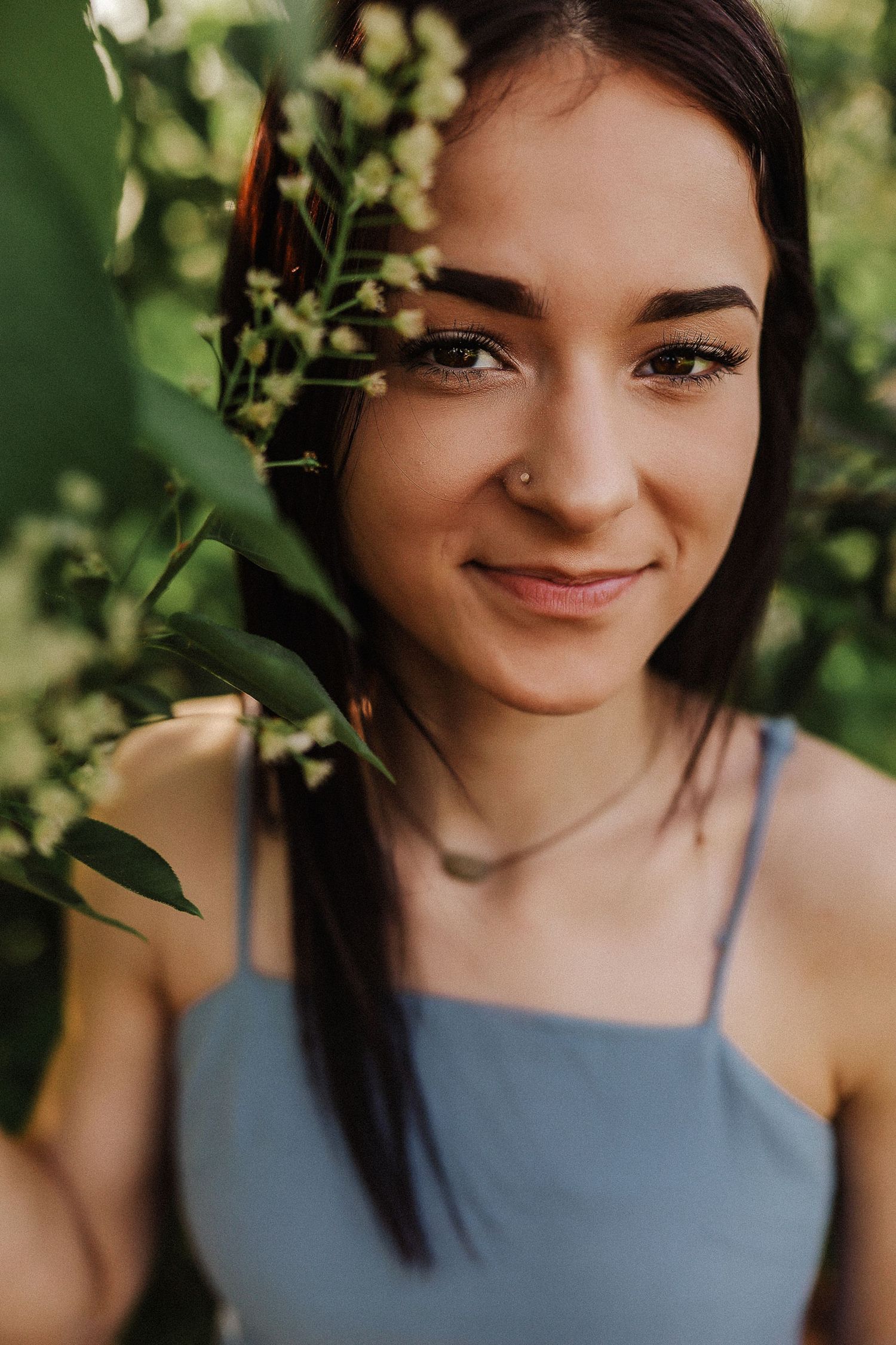 Portrait of a smiling woman with dark hair surrounded by green foliage in soft natural lighting.