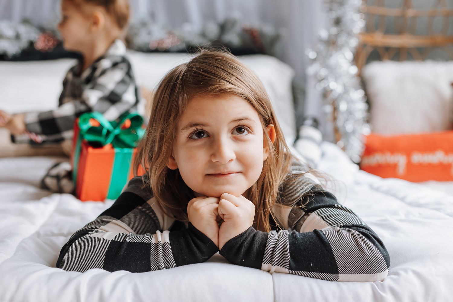 A child in plaid lying on white bedding during holiday season with presents in background.