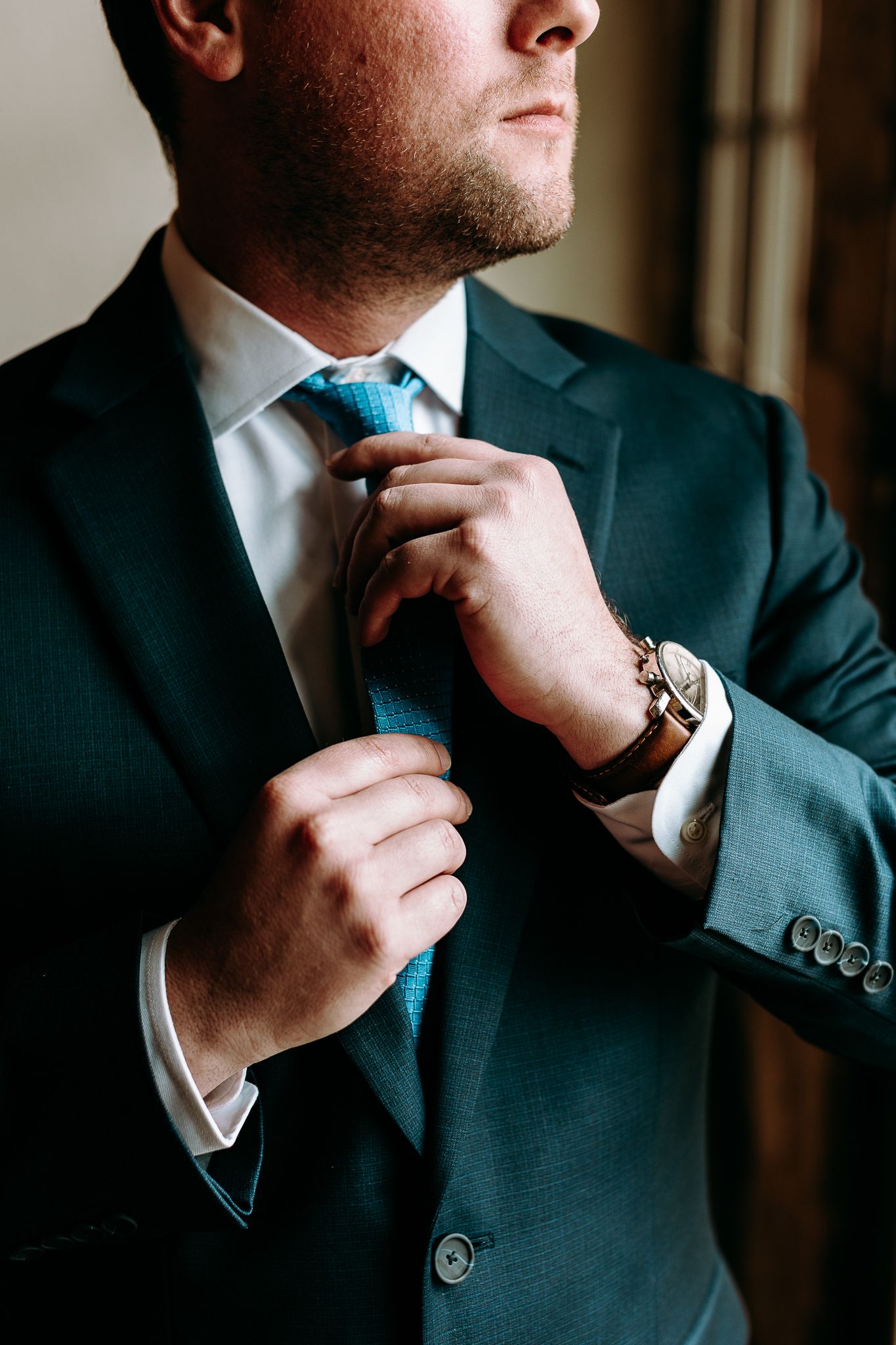 Close-up of man adjusting blue tie wearing dark suit and silver watch.