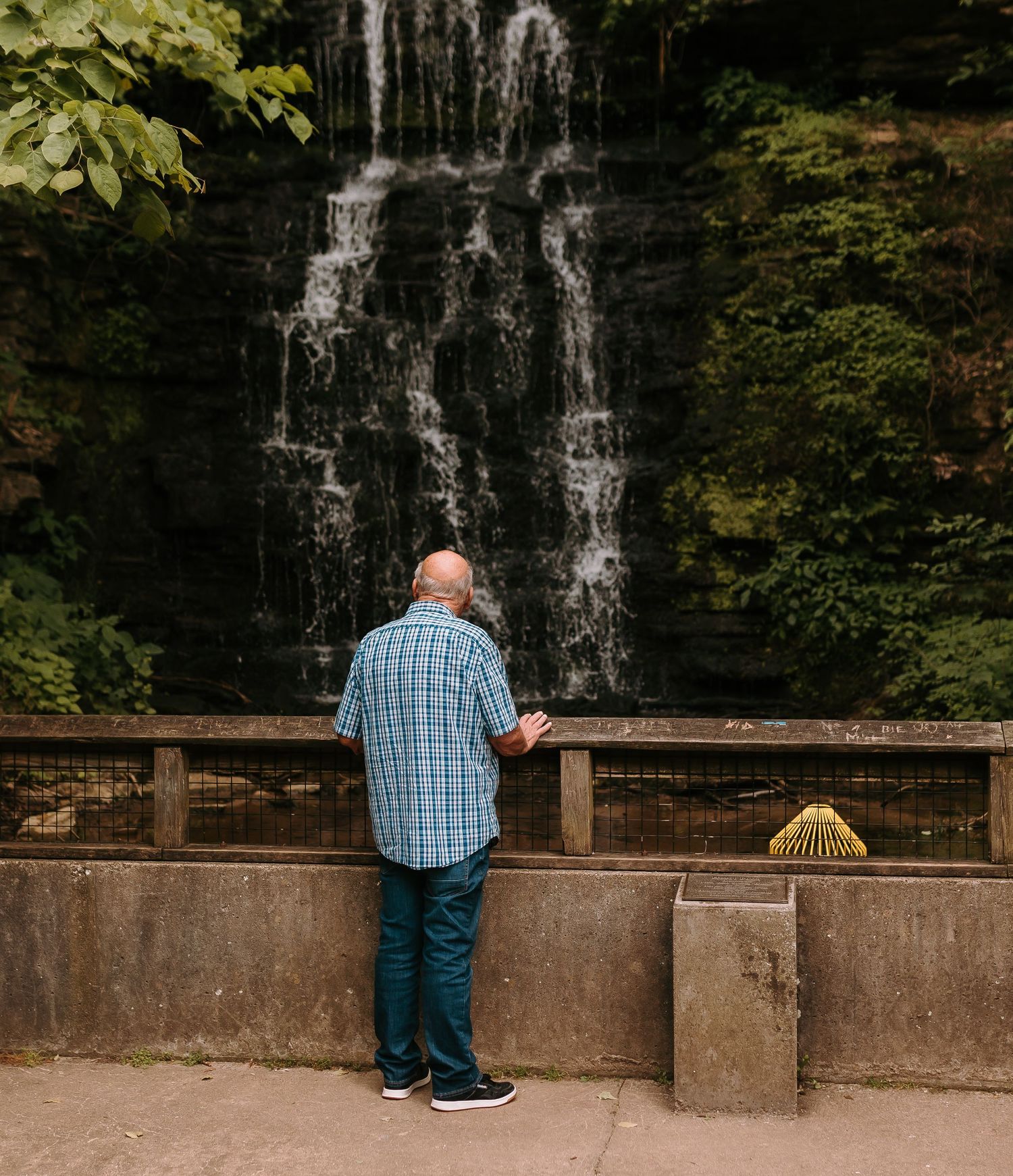 Bald man in blue plaid shirt stands at wooden railing viewing a cascading waterfall surrounded by lush greenery.