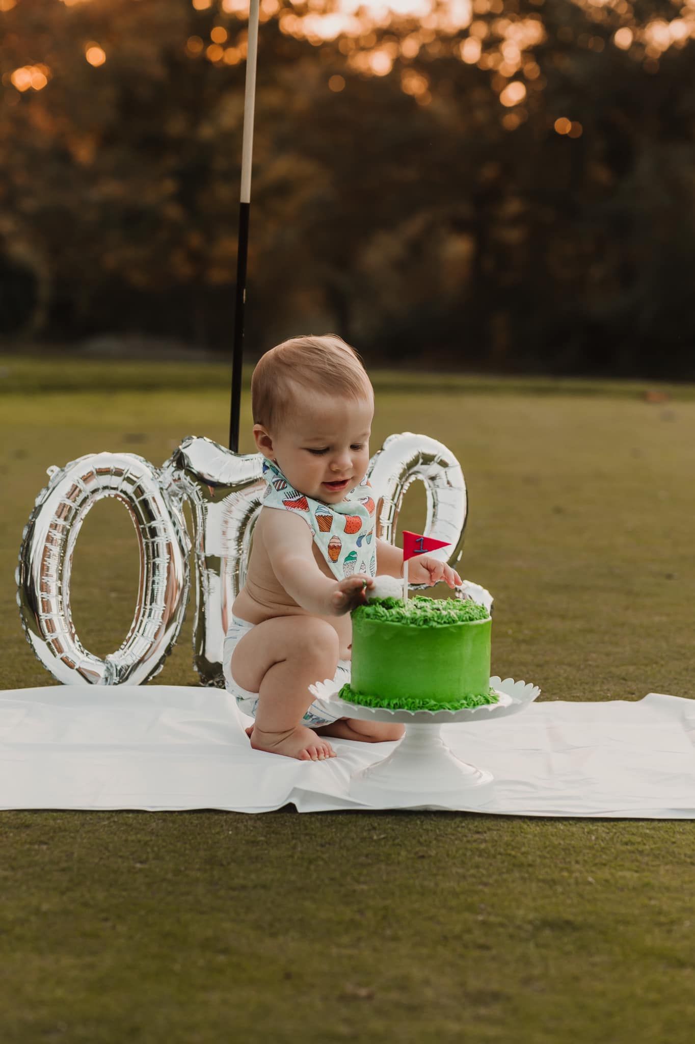 Little one explores their first birthday cake outdoors with silver balloon decorations at sunset.