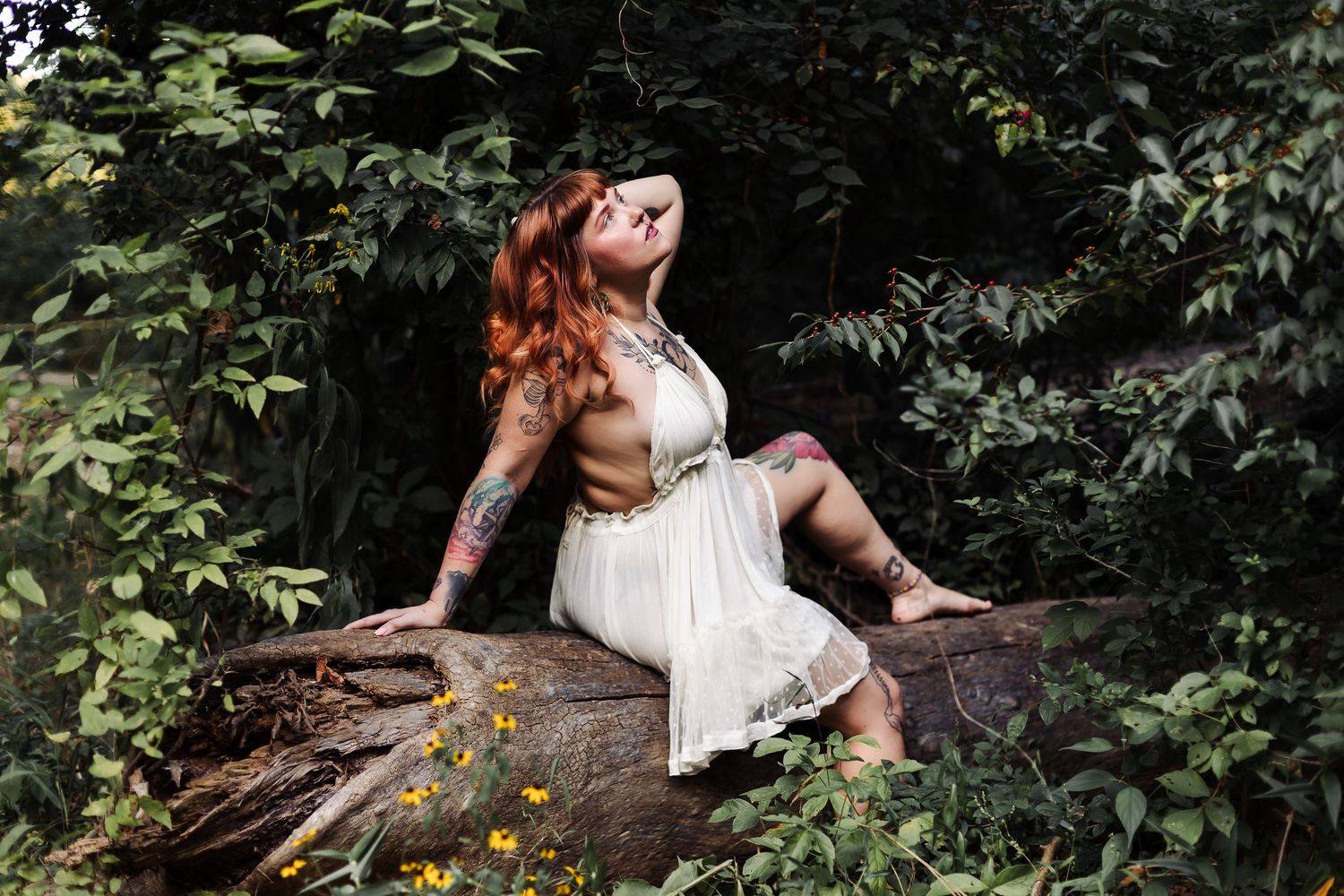 A person in a white dress poses dramatically on a fallen log in a lush forest setting with yellow wildflowers.