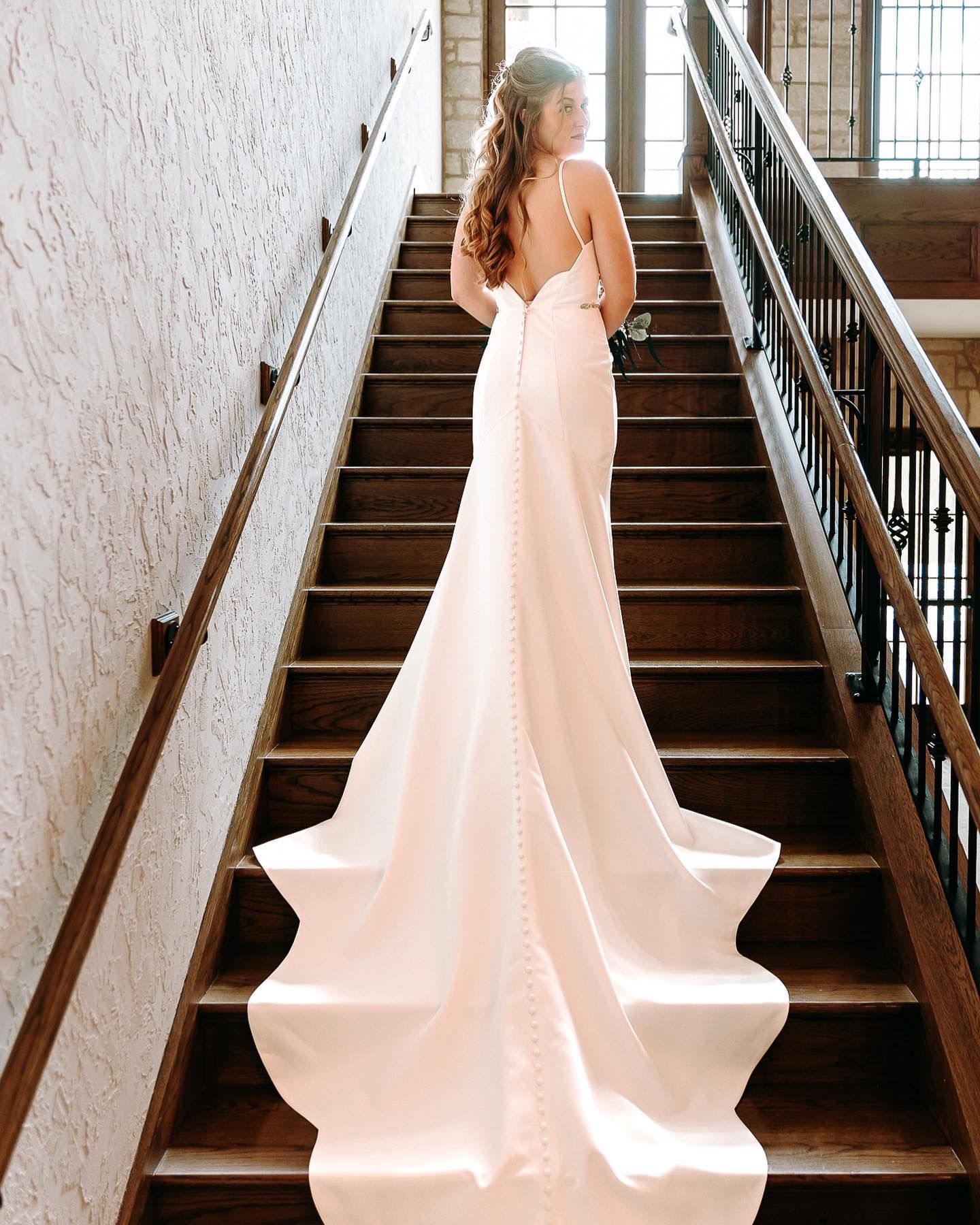 A blush pink wedding gown with a dramatic train cascades down wooden stairs.