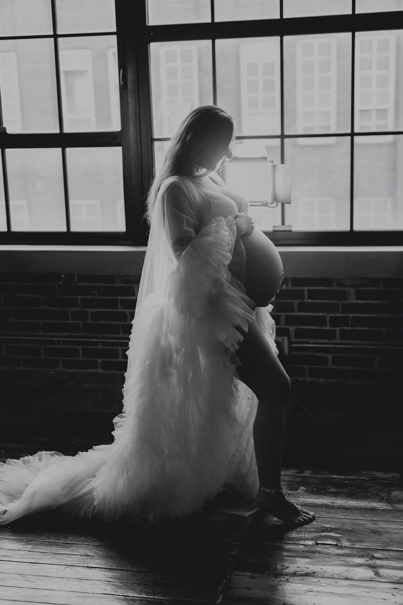 Black and white maternity photo of pregnant woman in flowing white gown standing by large windows in rustic loft.