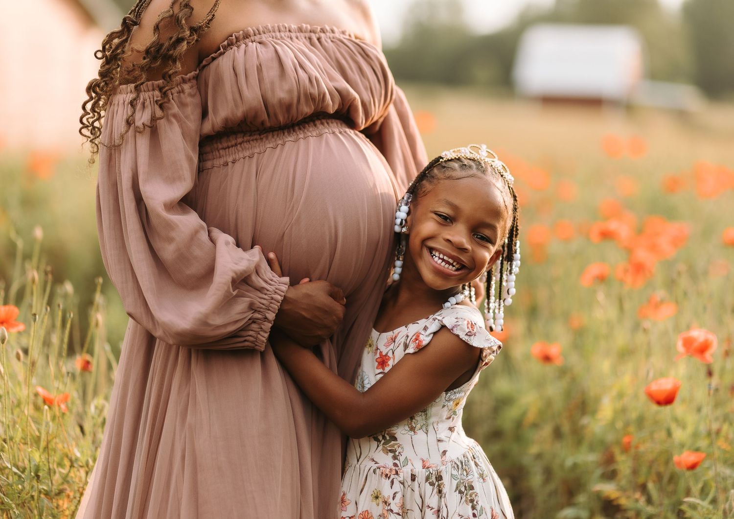 A young girl hugs her pregnant mother in a flower field, both wearing dresses, smiling joyfully outdoors.