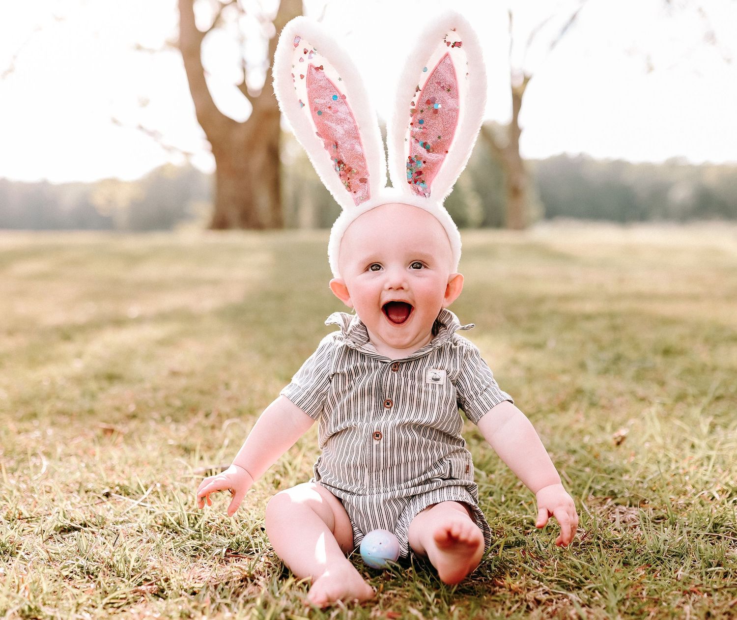A baby wearing pink bunny ears and striped outfit sits laughing on grass.