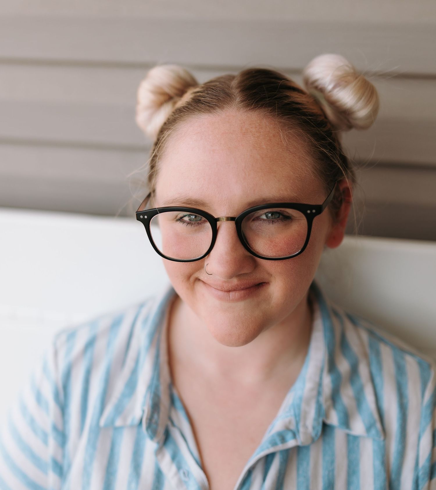 A casual portrait of someone with blonde hair buns and glasses against a light wall.