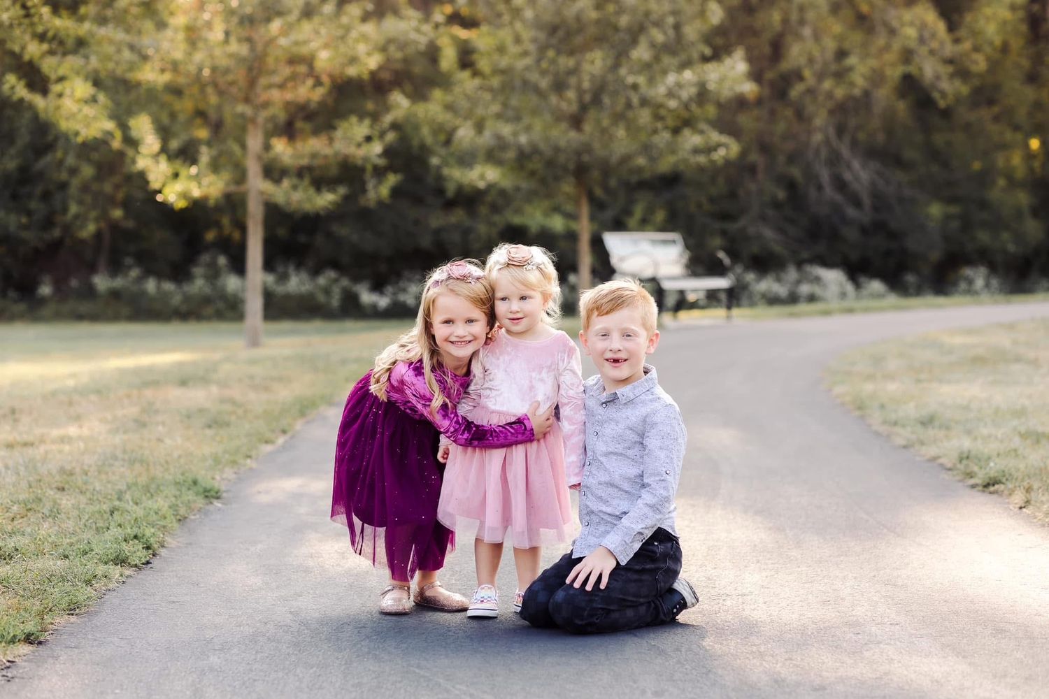 Three small children in formal attire smile together while posing on a walking path in an outdoor setting.
