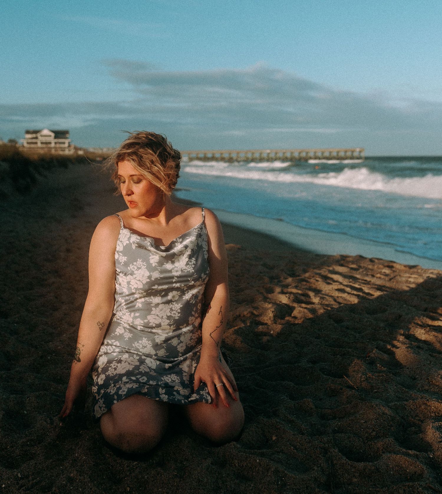 Woman in floral dress kneeling on sandy beach at golden hour with ocean waves and pier in background.