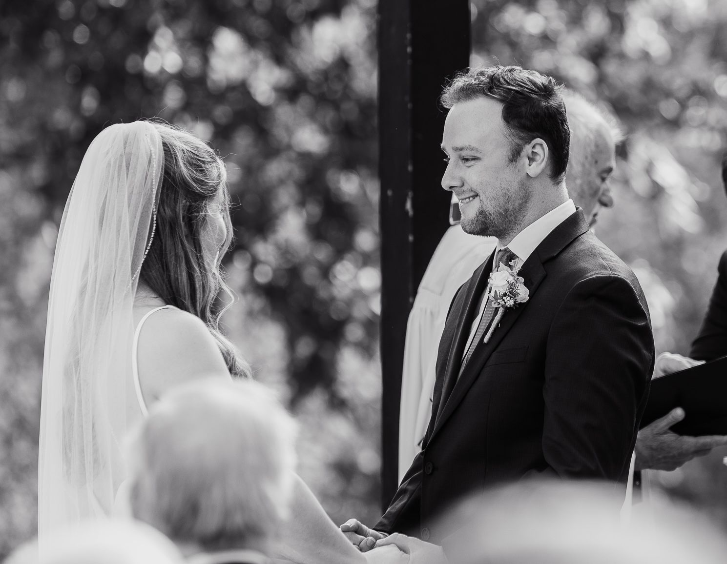 Black and white ceremony photo of couple exchanging vows outdoors.