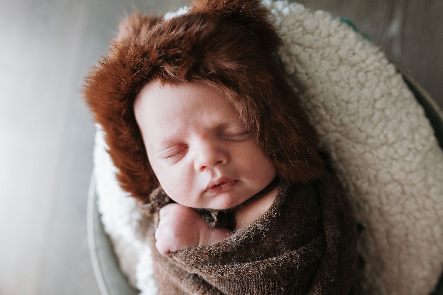 Newborn baby with red hair sleeping peacefully in brown swaddle and white basket.