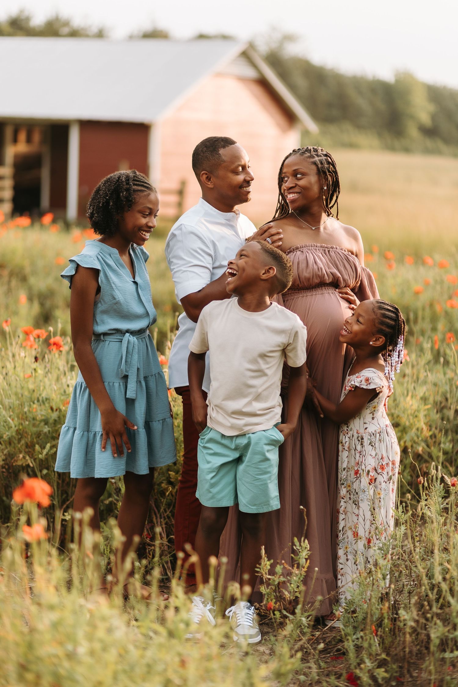 A joyful Black family poses in a flower field, with a pregnant mother, father, and three children near a red barn.