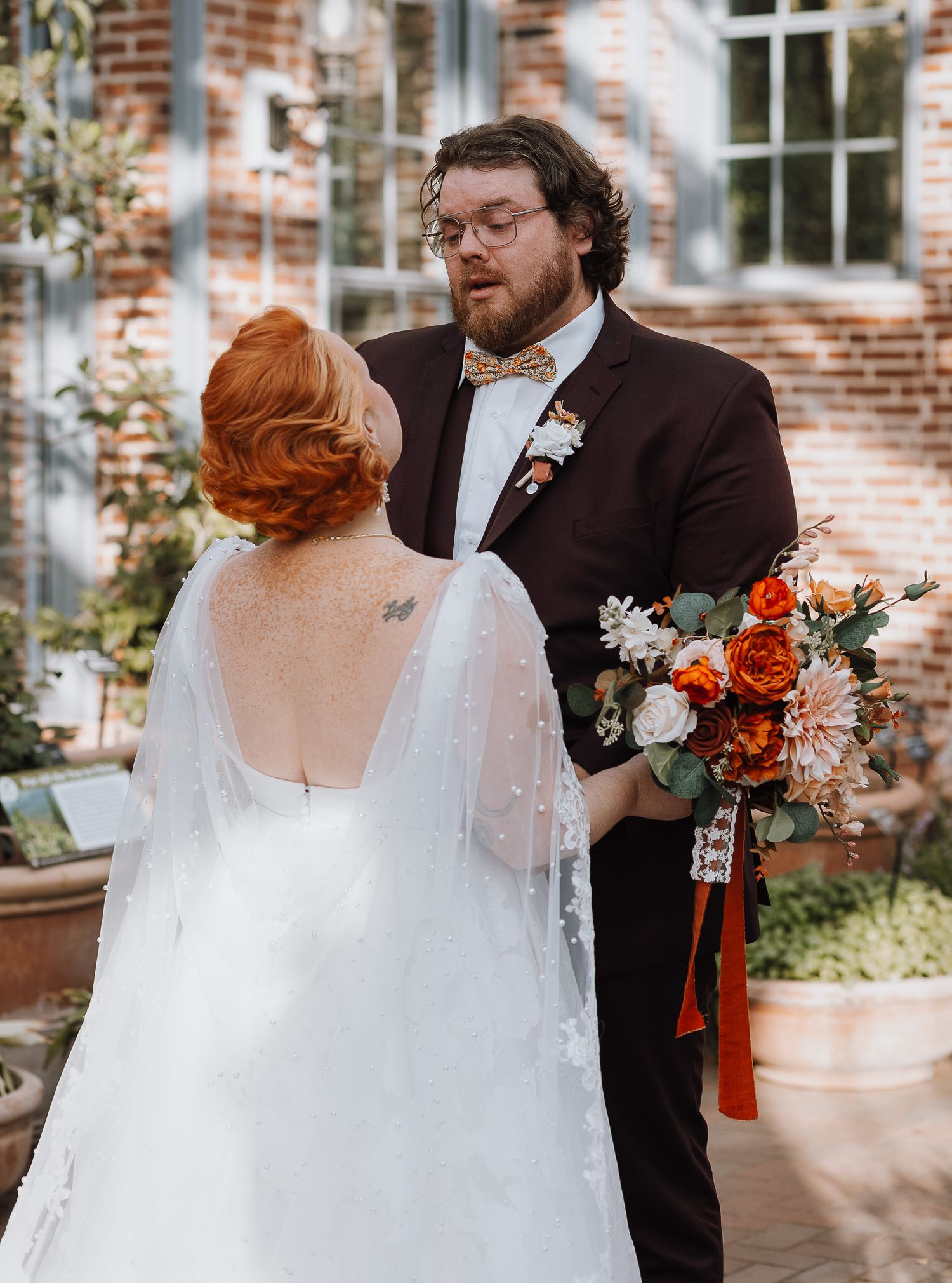 Couple in wedding attire share tender moment against brick building backdrop.