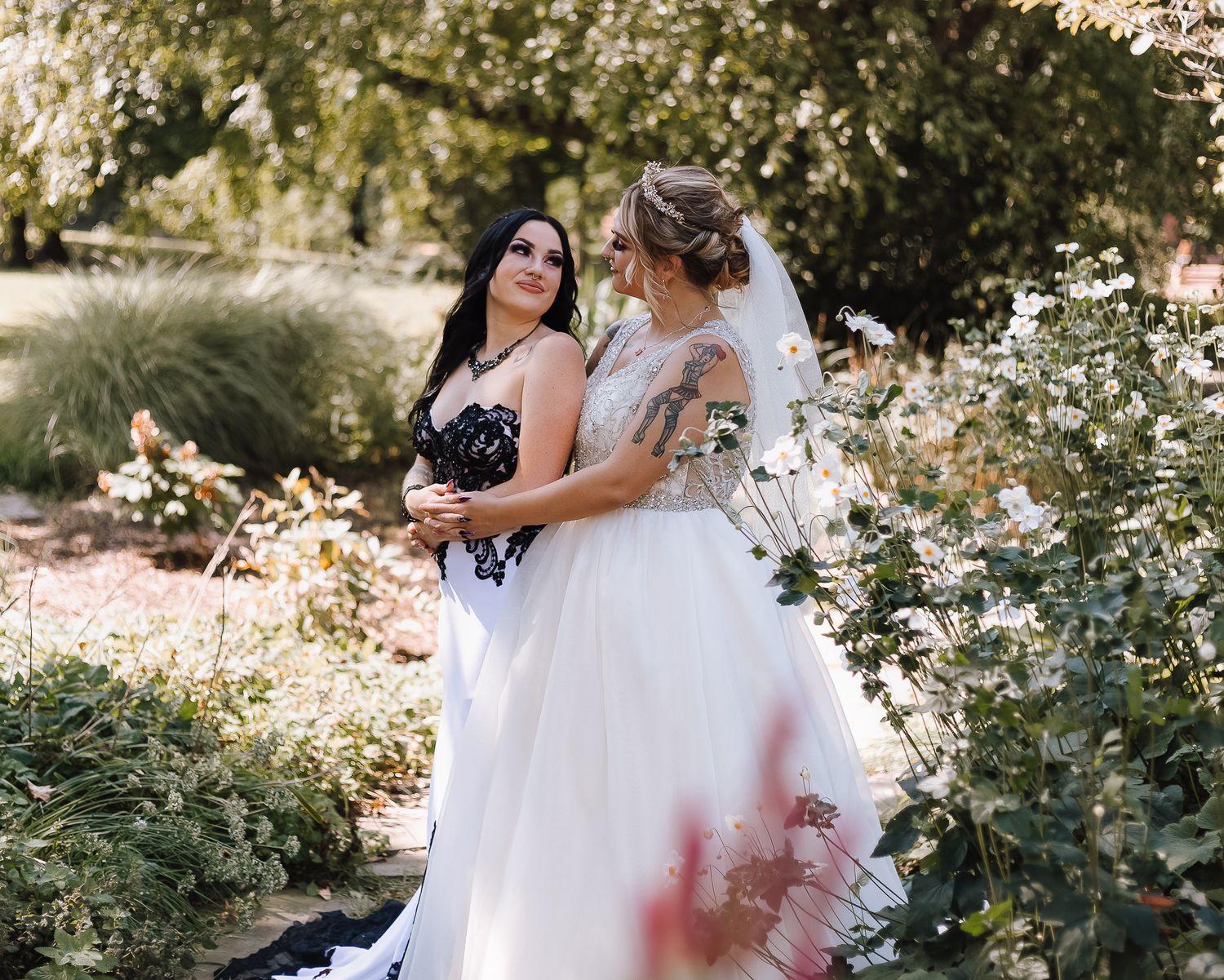 Two brides in elegant gowns embrace in a sunlit garden setting with blooming flowers.