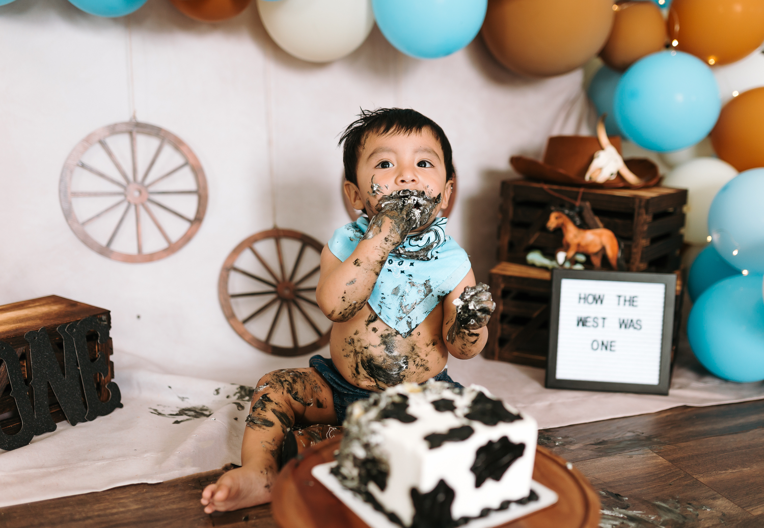 Baby enjoying a cow print cake smash with western balloon backdrop and wagon wheel decorations.