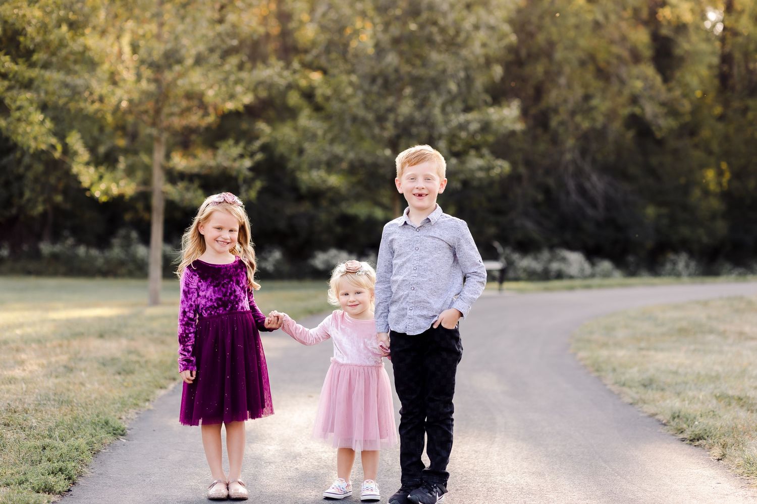 Three young children pose on a park path, two girls in pink and purple dresses and a boy in gray shirt and black pants.