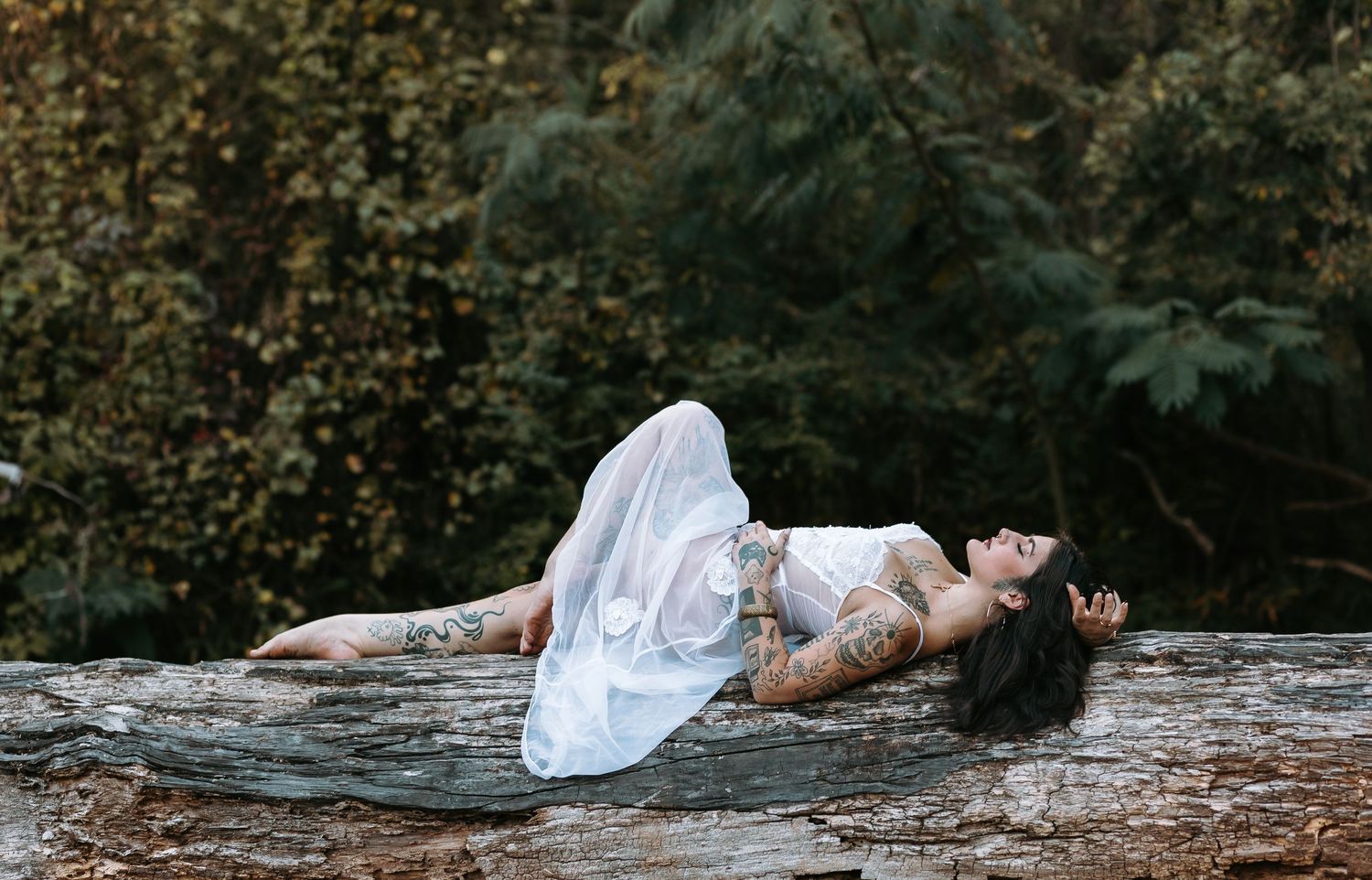 A tattooed woman in a white dress reclines gracefully on a large rock surrounded by lush green forest trees.