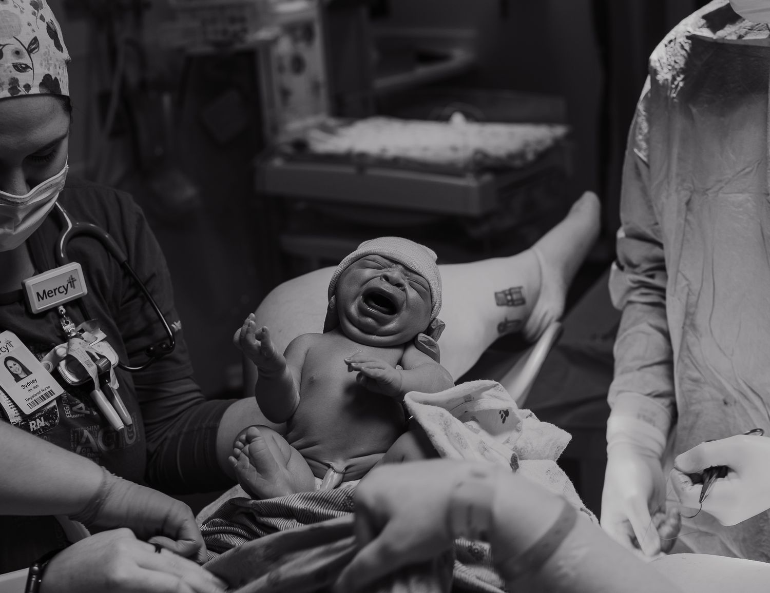 Black and white photo of a newborn baby crying moments after birth, held by medical staff in a hospital delivery room.
