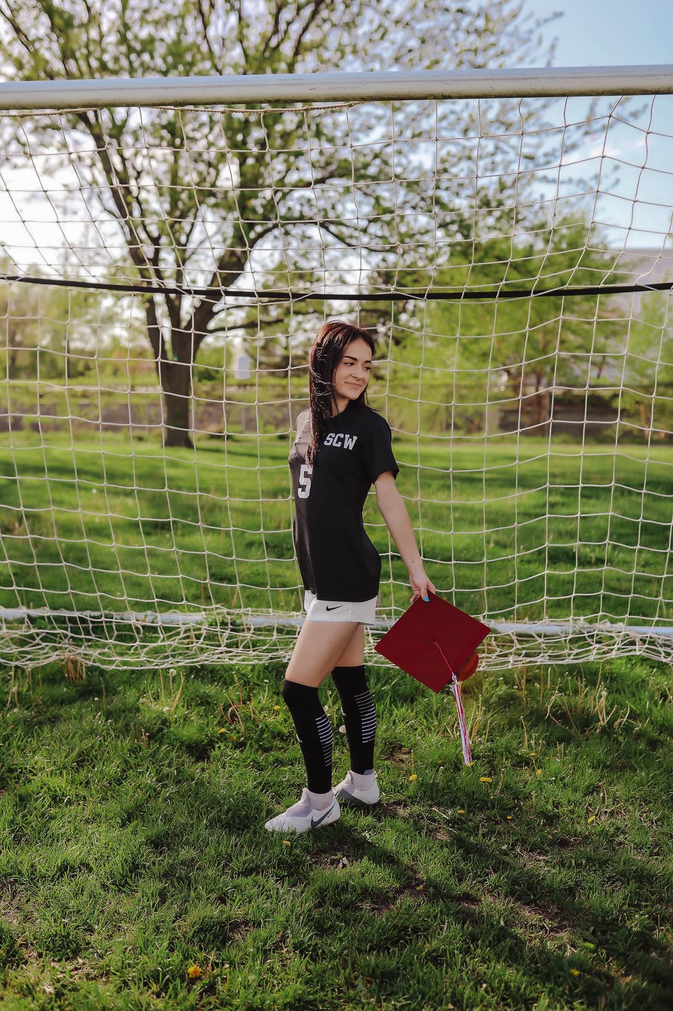 Senior graduation photo at soccer goal wearing black jersey and holding red book.