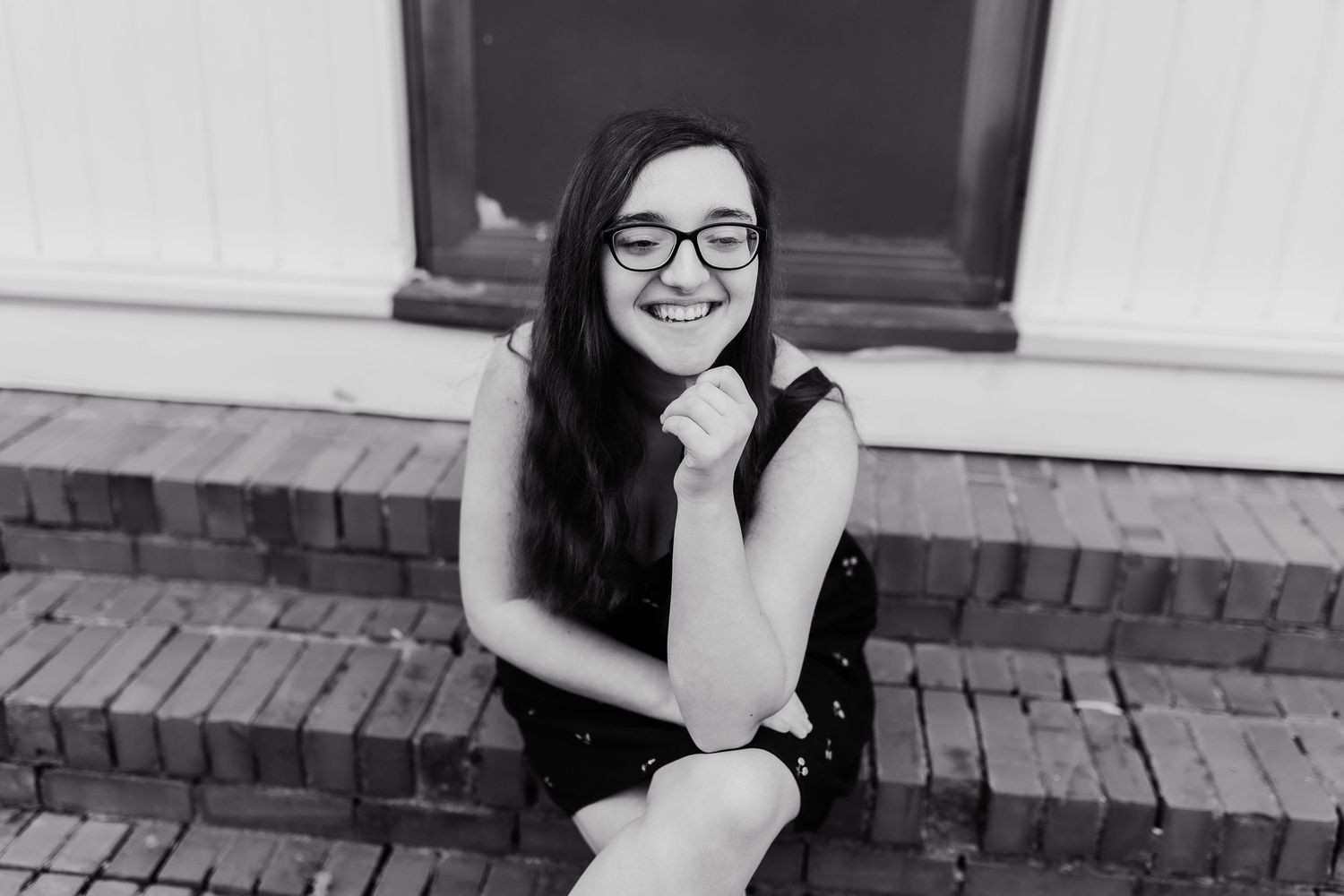 Young woman with glasses and long dark hair smiling on brick steps in black and white photo.