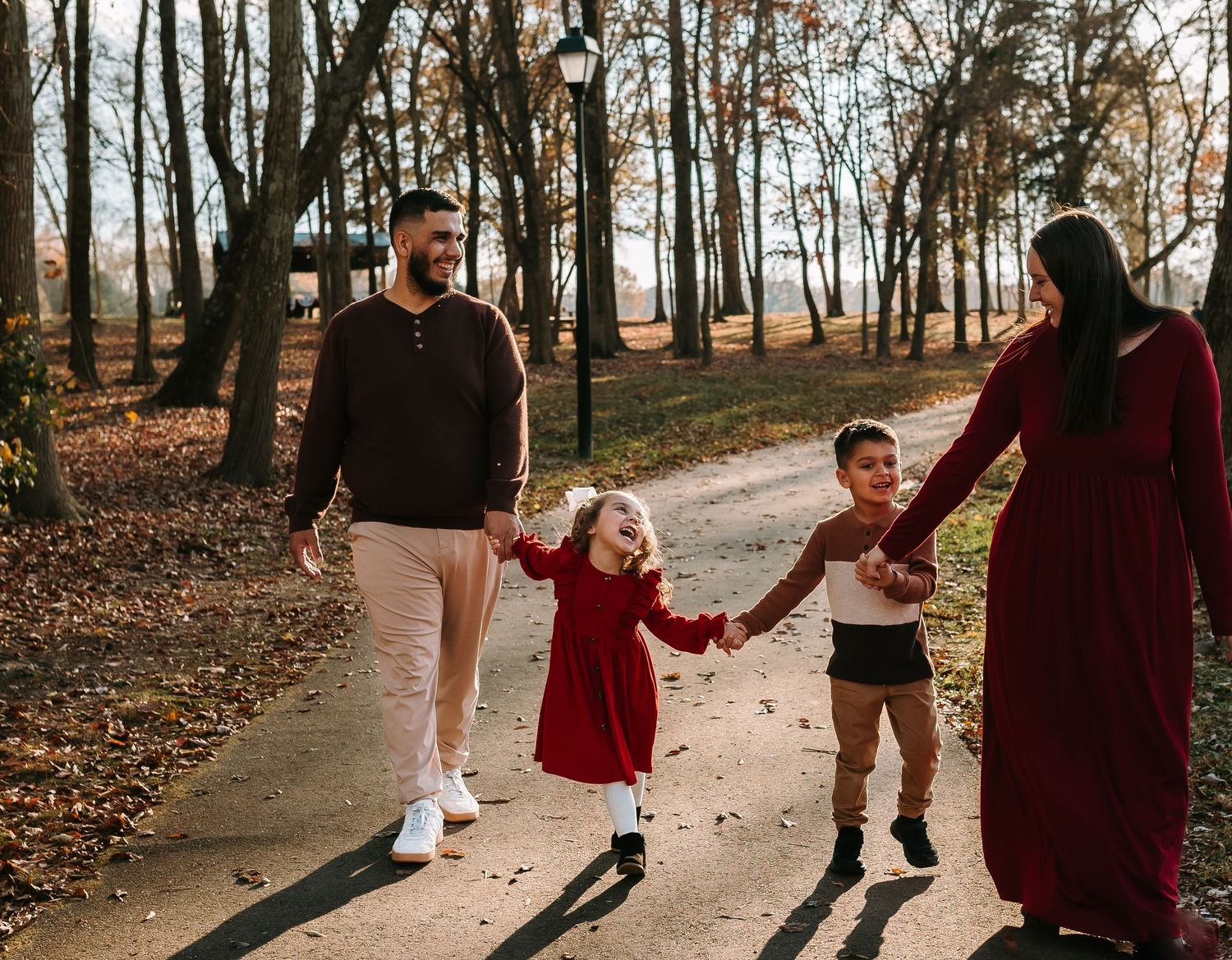 Family of four walking hand-in-hand on a park path surrounded by autumn trees, wearing coordinated red and neutral outfits.