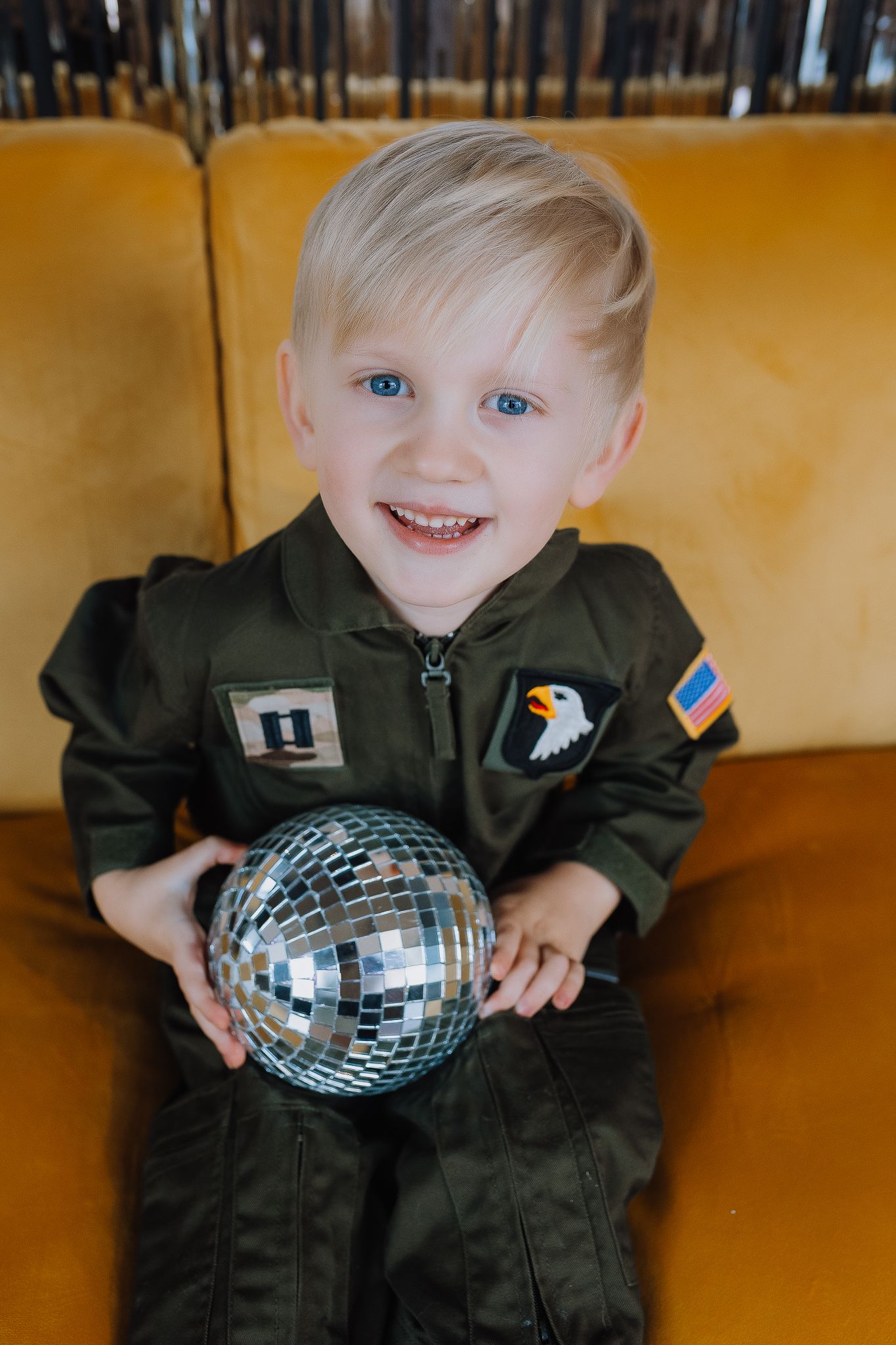 Smiling toddler in green military pilot costume holding a disco ball, sitting on a yellow sofa.