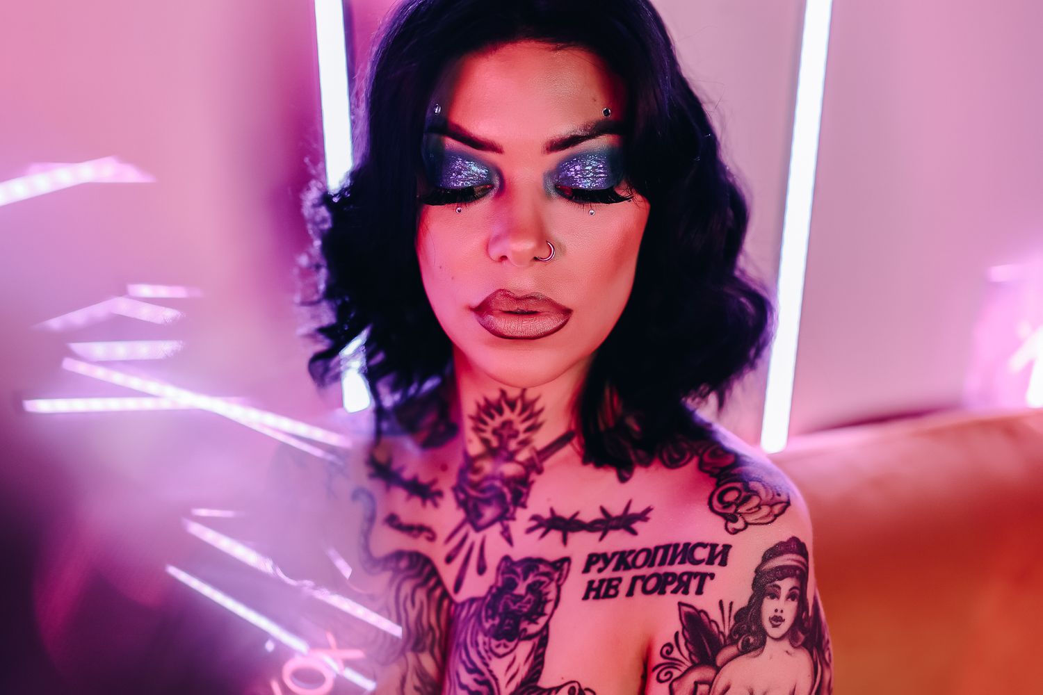 Tattooed woman with bold blue eyeshadow and neck tattoos poses under pink and purple neon lights.