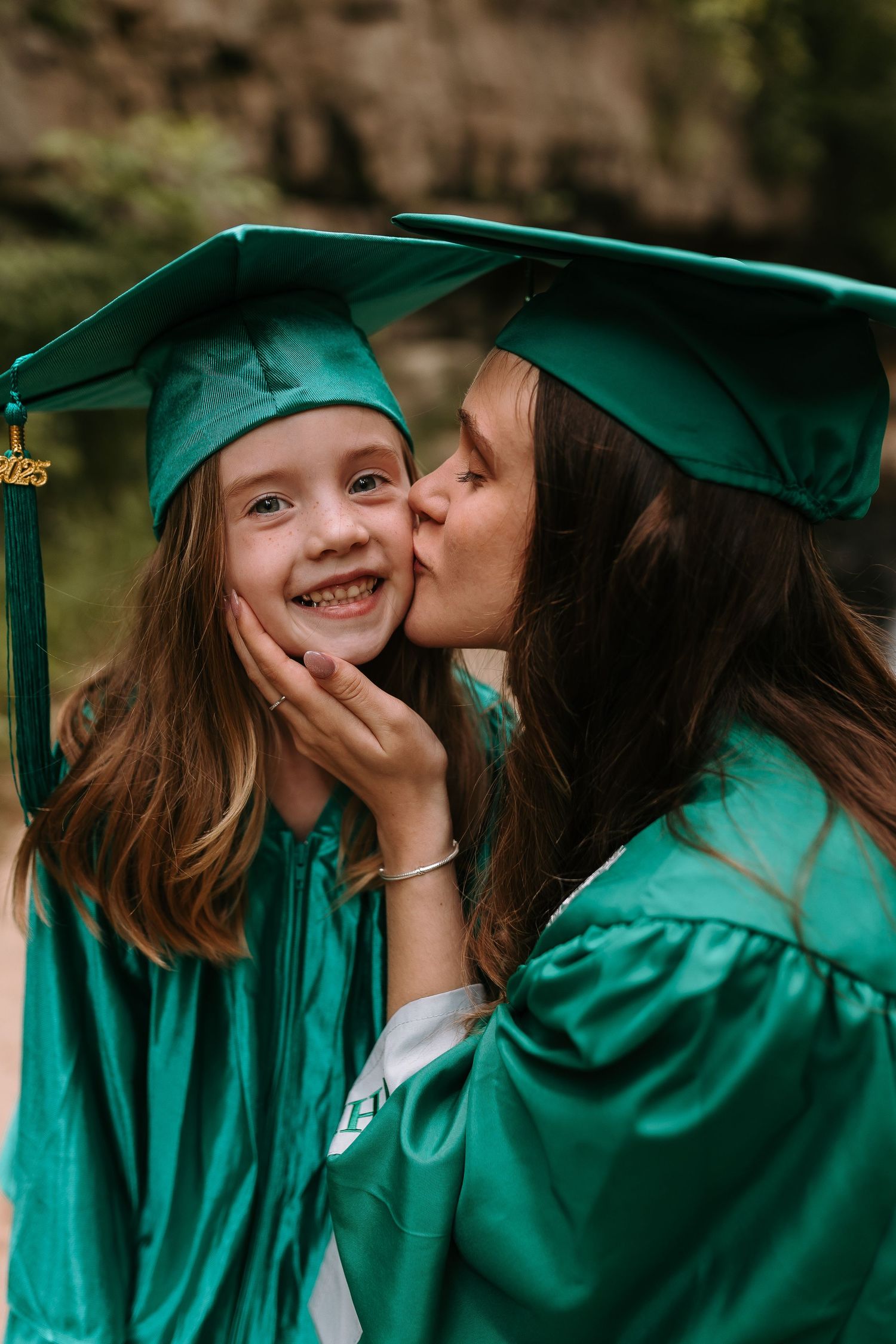 Two girls in green graduation caps and gowns share a sweet kiss on the cheek during a graduation celebration.