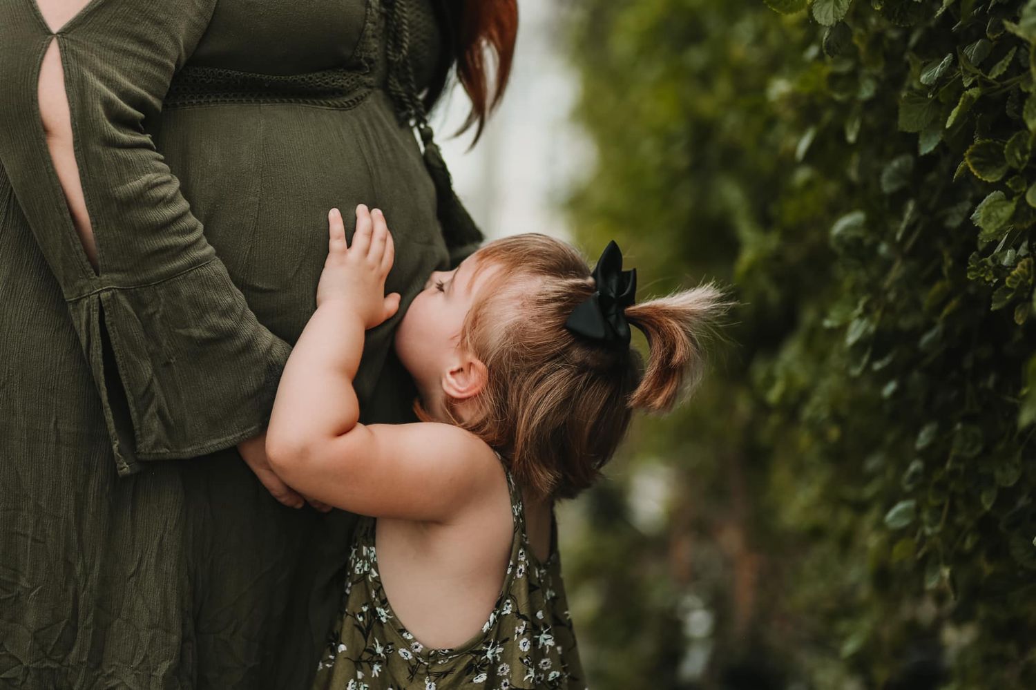 A young child in floral dress touching a pregnant belly in an outdoor garden setting.