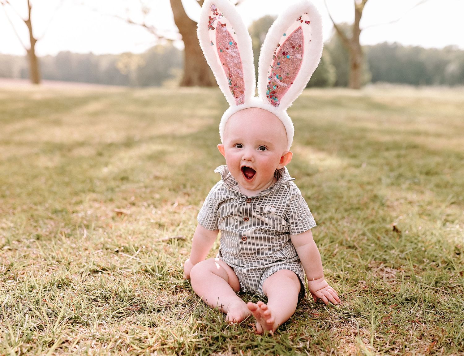 Baby wearing bunny ears and striped outfit sitting on grass with excited expression.