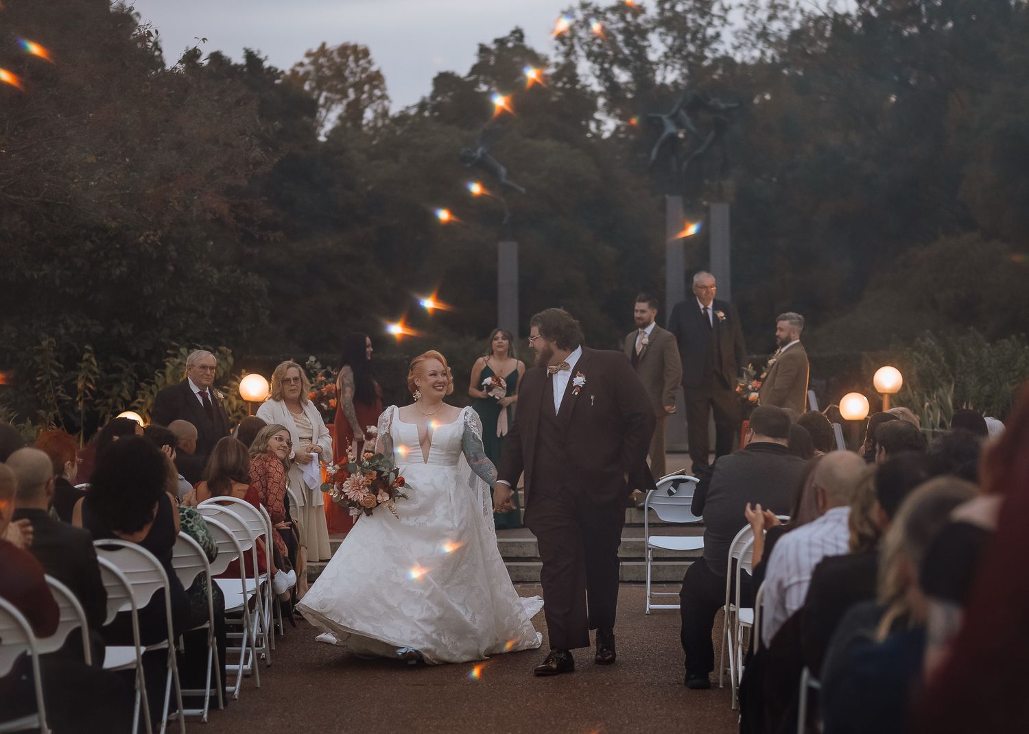 Couple walking down aisle at twilight outdoor wedding ceremony with string lights overhead.