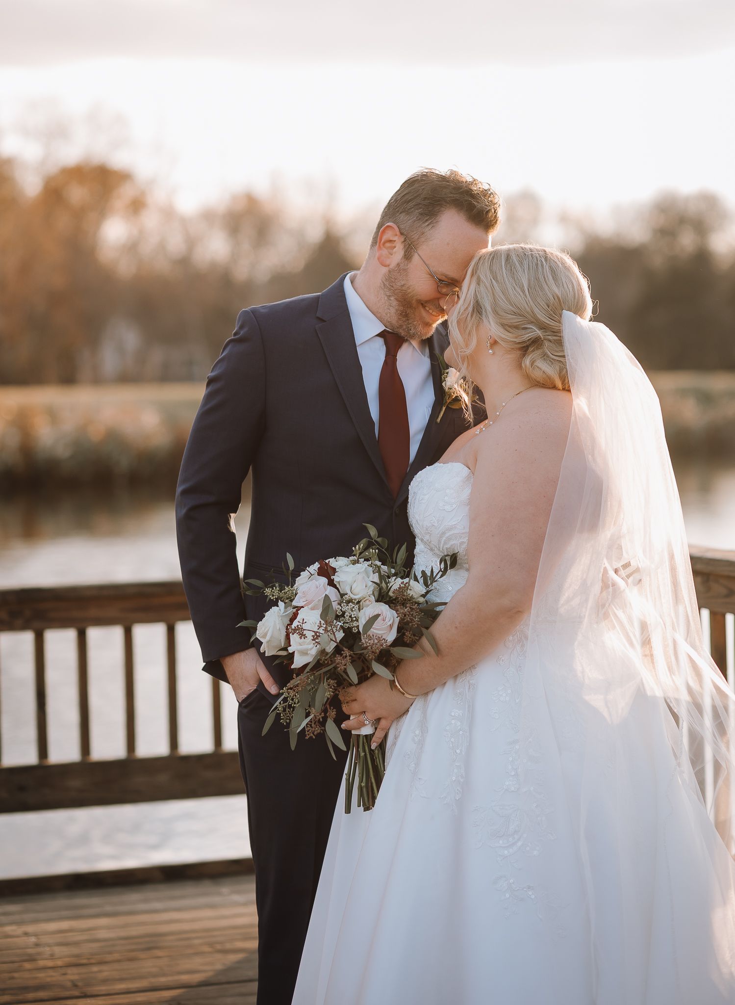 Newlyweds share an intimate moment on a wooden deck during golden hour with a bouquet of white flowers.