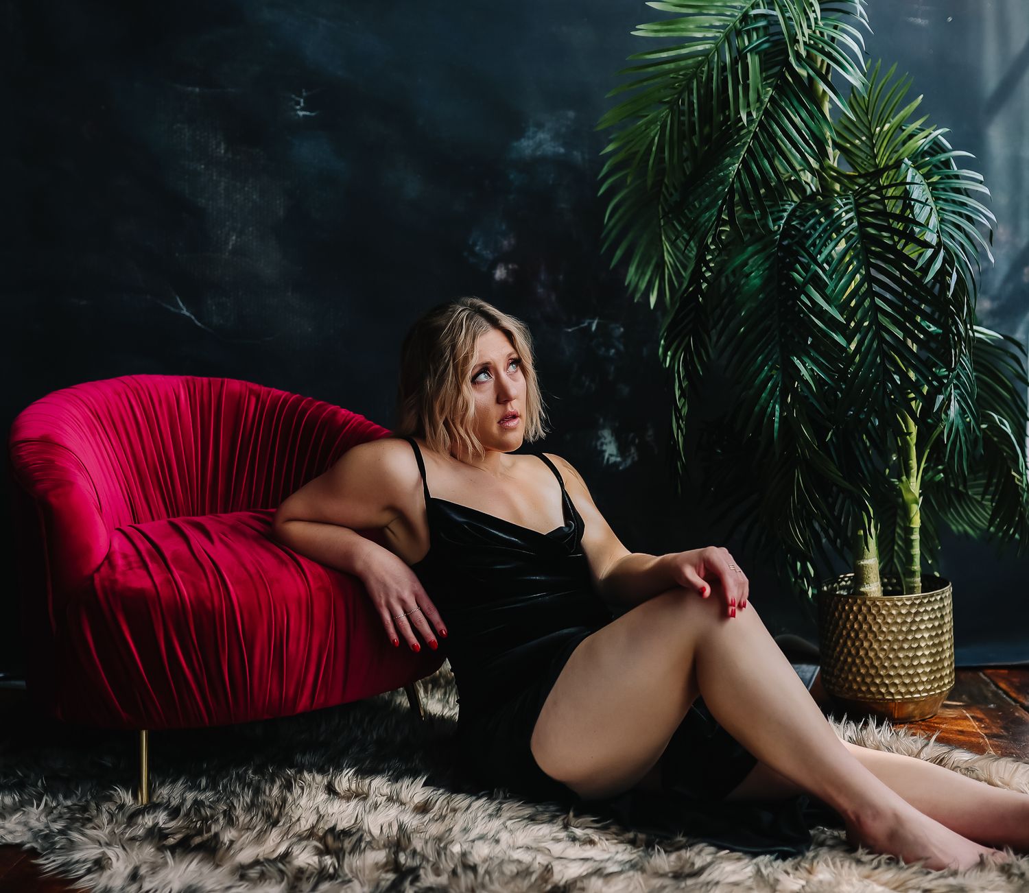 Woman in black dress sitting on fur rug beside red velvet chair and gold potted palm plant.