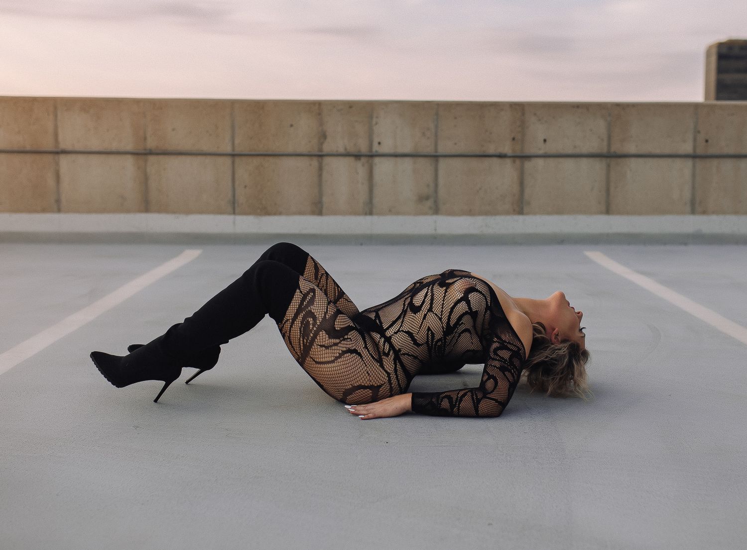 Woman in black lace bodysuit and thigh-high boots posing on rooftop parking lot.