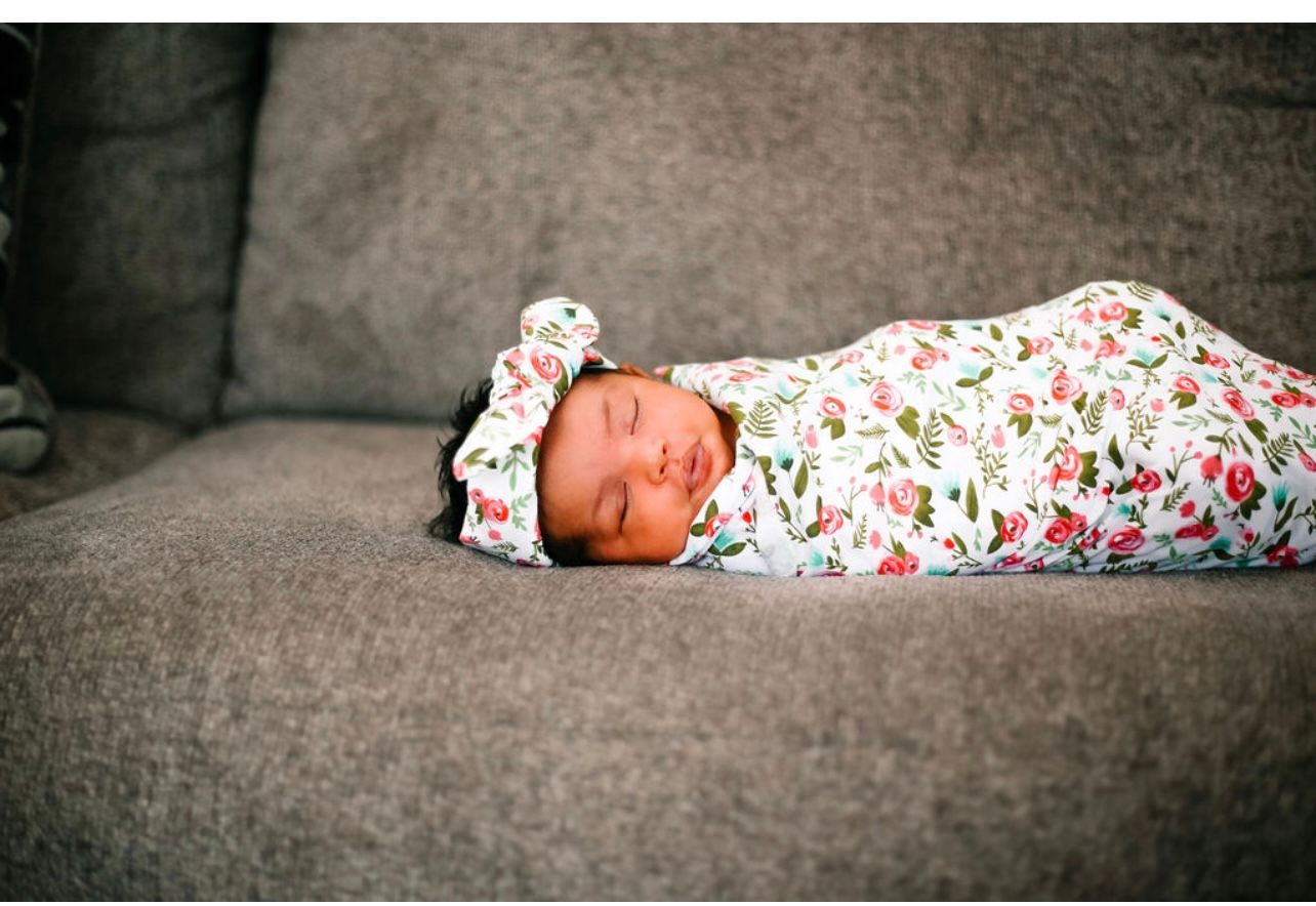 A sleeping infant swaddled in floral print fabric rests on a gray couch.