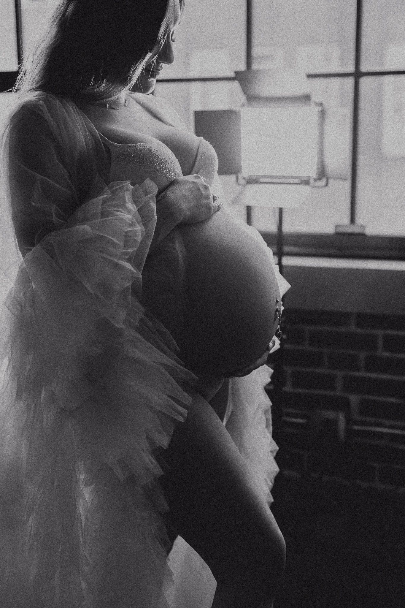 Black and white maternity photo of pregnant woman in lace gown by window.