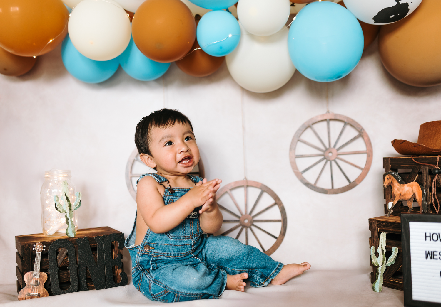 Baby boy in denim overalls sits smiling at a western-themed first birthday photo shoot with colorful balloons.