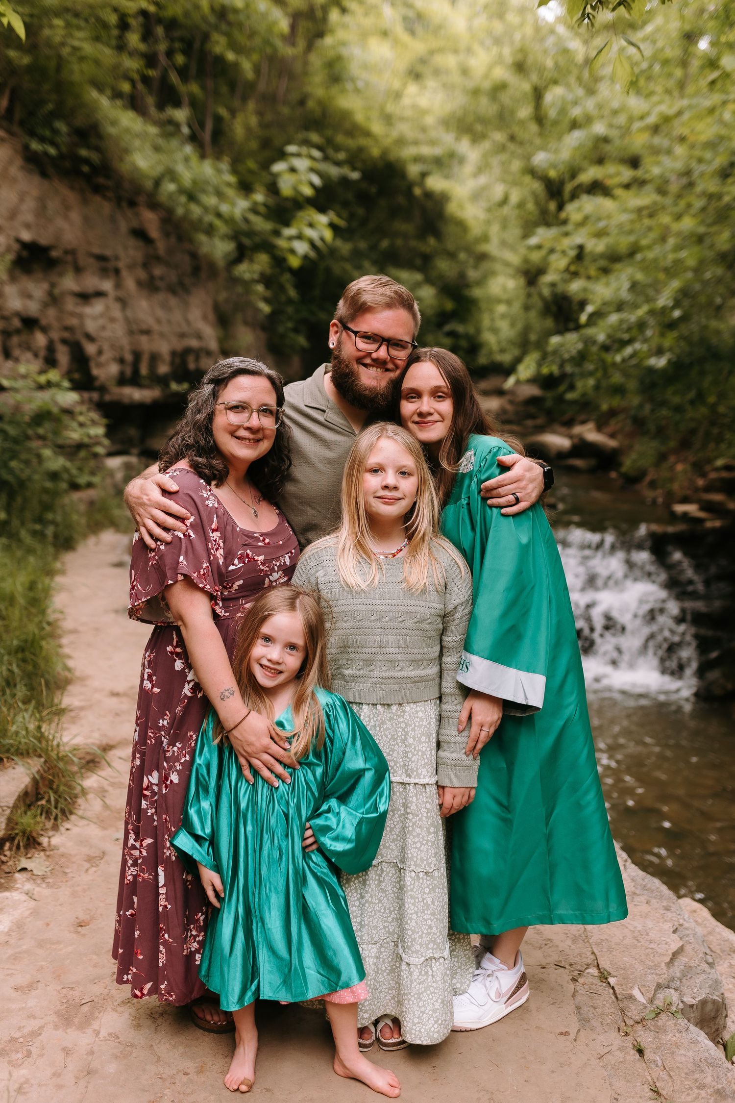 Family of five posing outdoors near a waterfall, with two girls in teal dresses and a graduate in green gown.