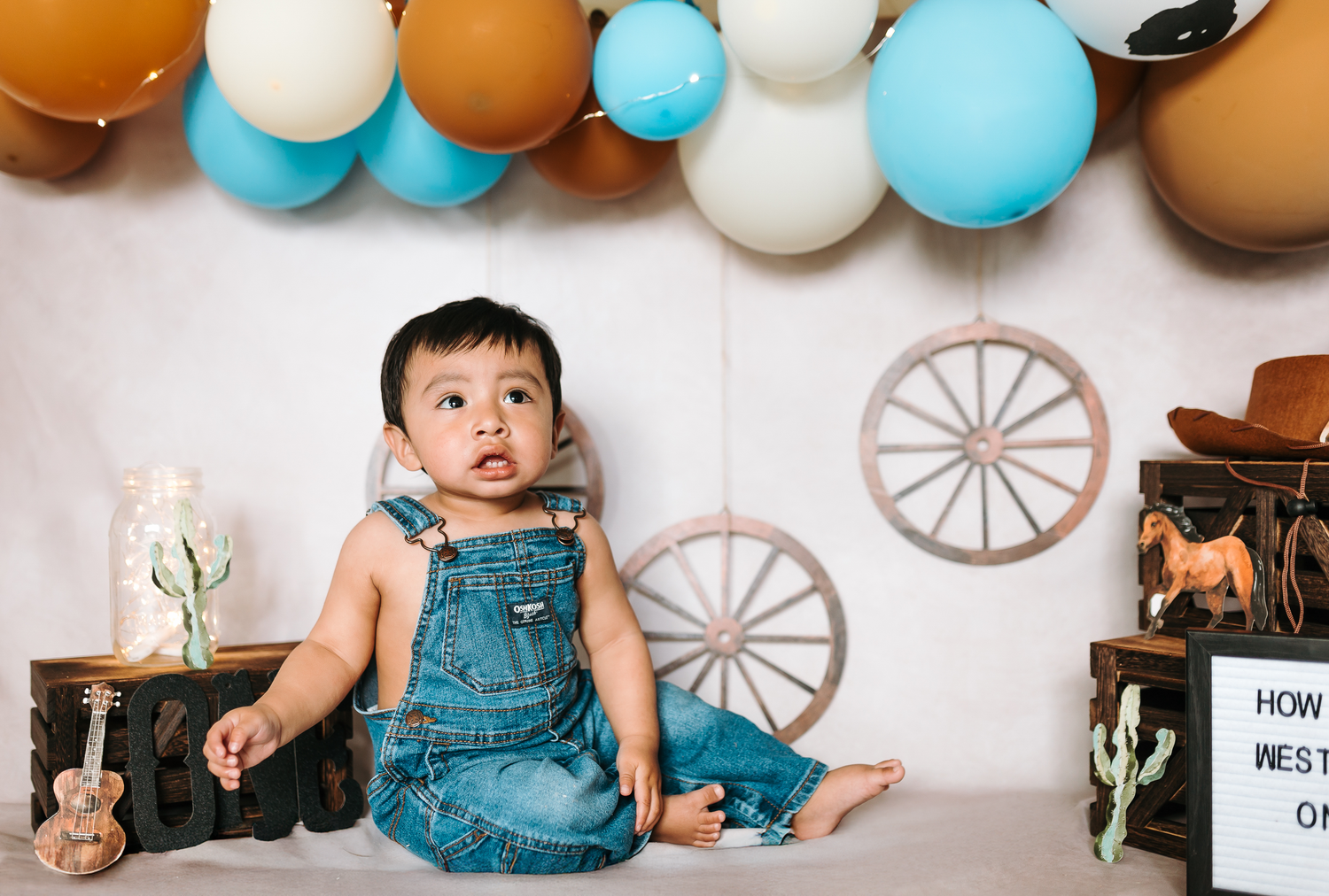 Cowboy-themed birthday photo with balloon arch and rustic decorations featuring a child in overalls.