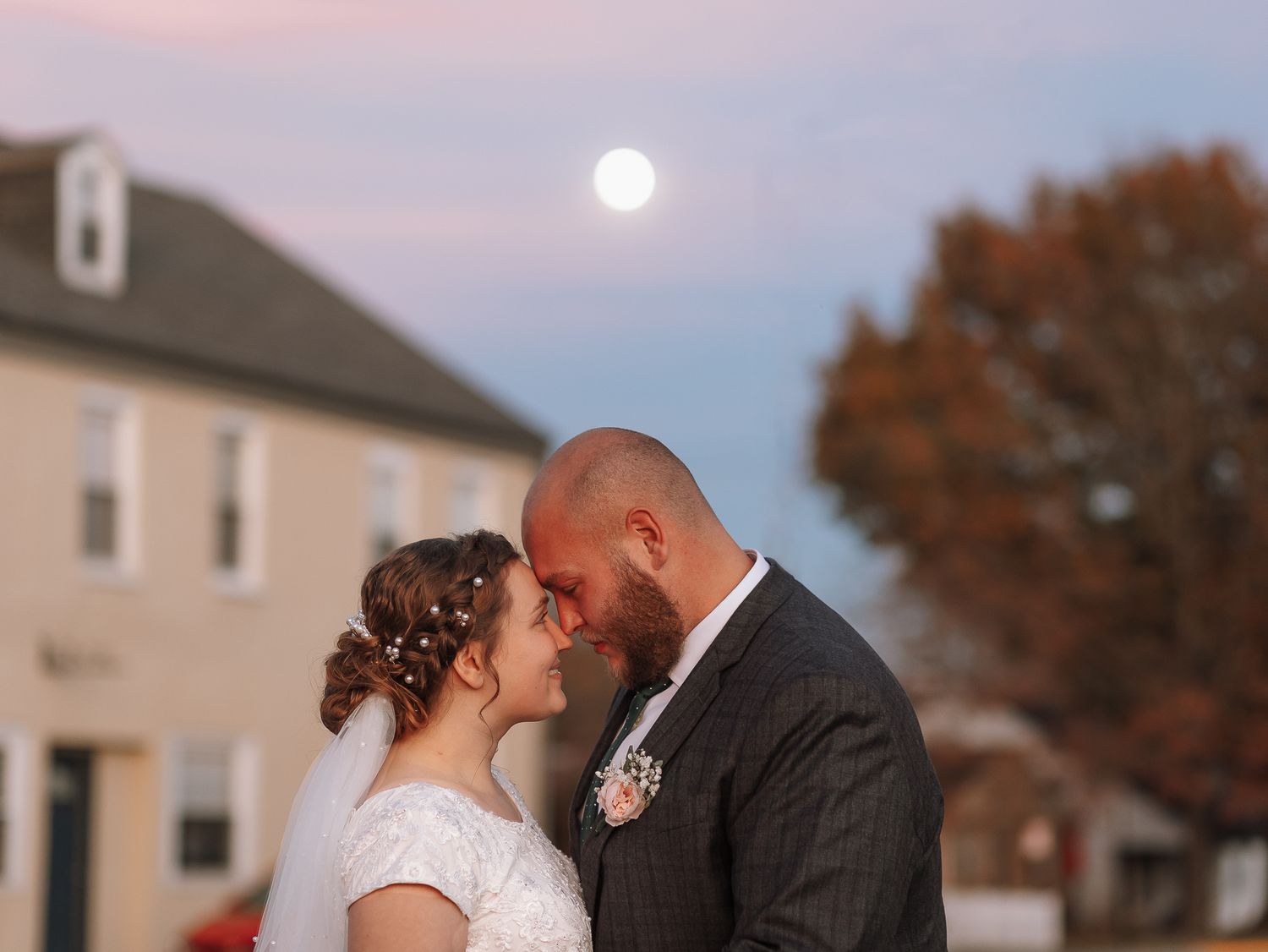 Tender moment between newlyweds at dusk with full moon visible.